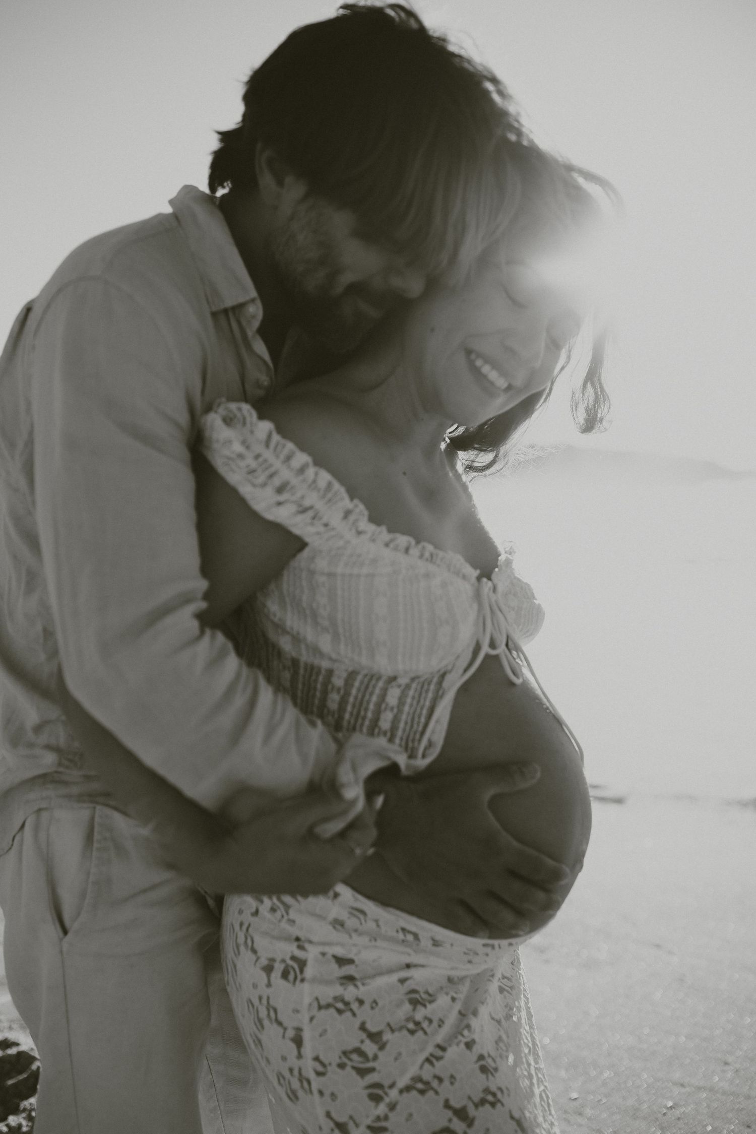 Black and white photo of tender embrace between couple, woman in white lace dress showing pregnant belly.