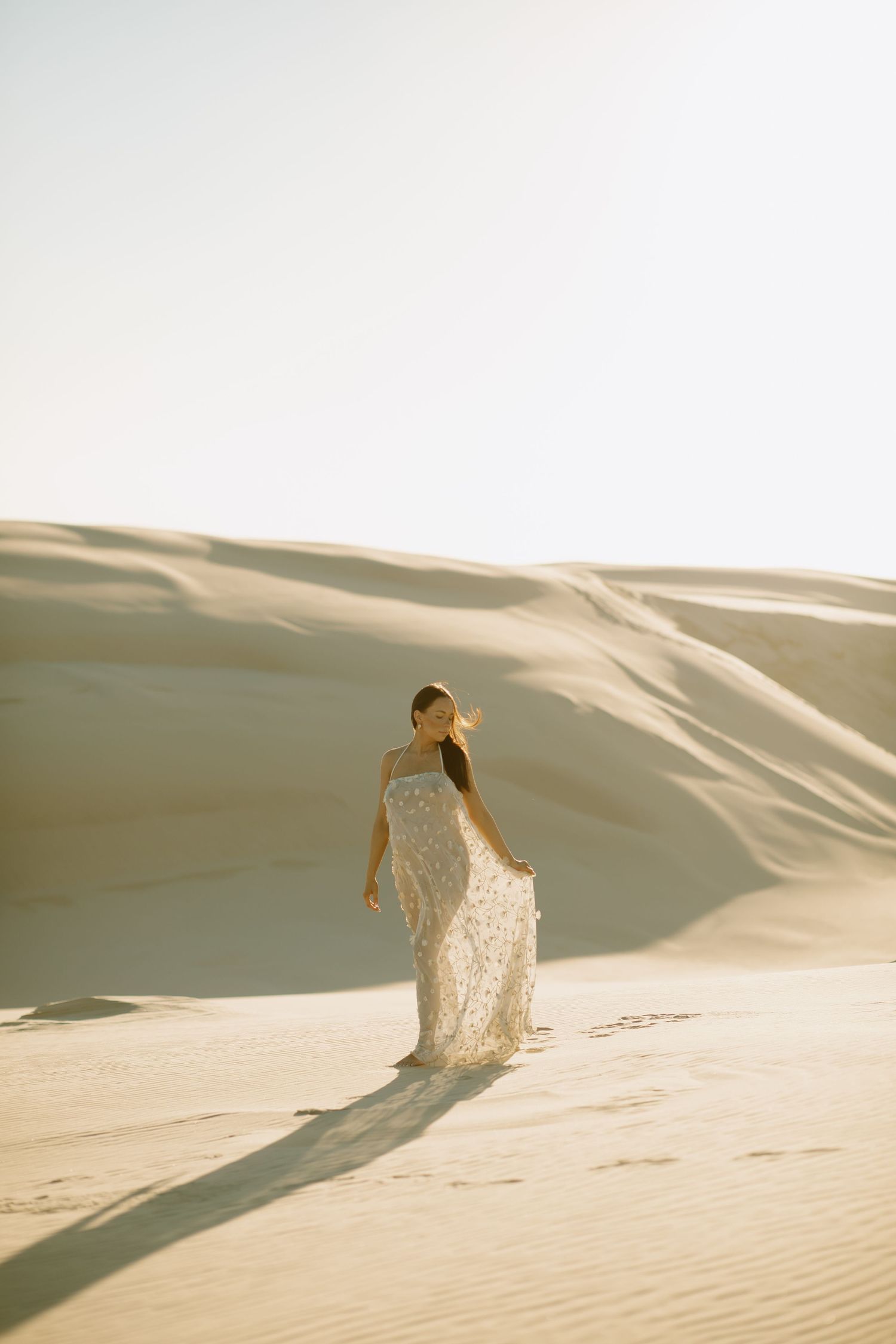 A person in a flowing white lace dress poses gracefully on rolling sand dunes during golden hour.