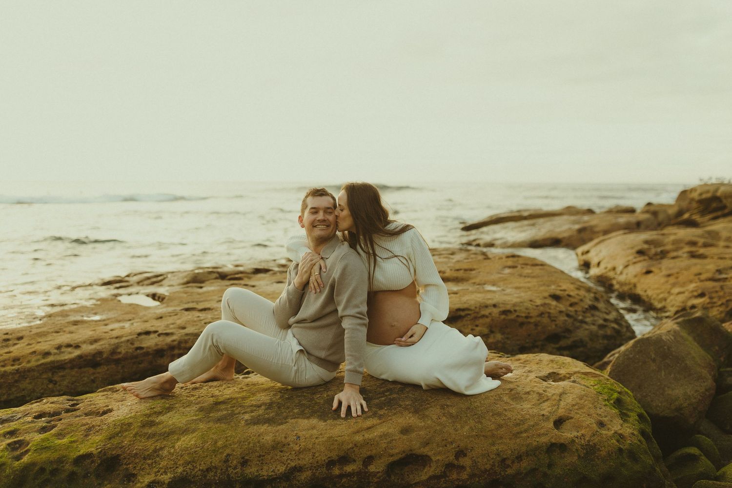 A couple sits together on coastal rocks at sunset overlooking the ocean on a misty day.