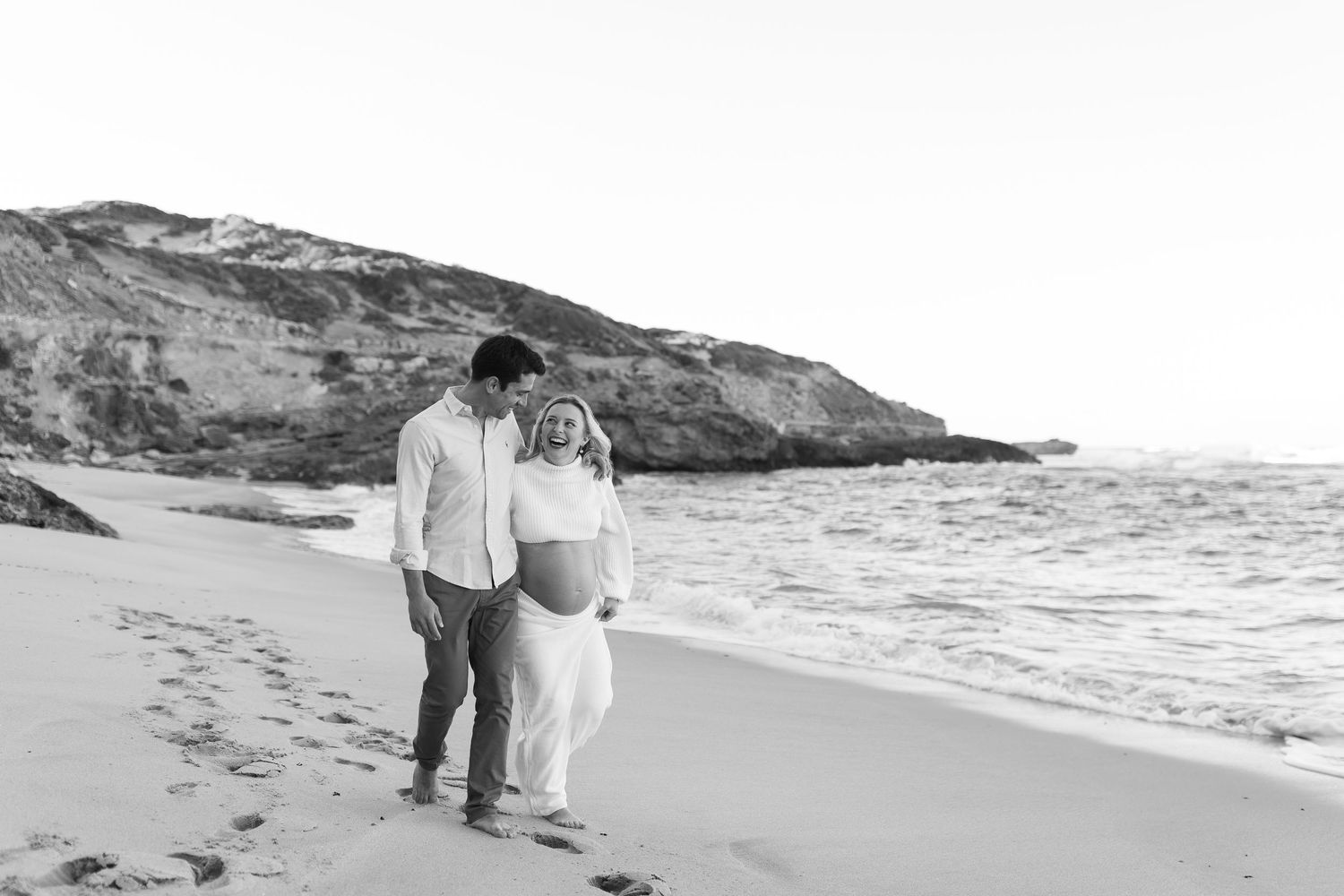 A couple walks together along a scenic beach shoreline with rocky cliffs in the background in black and white.