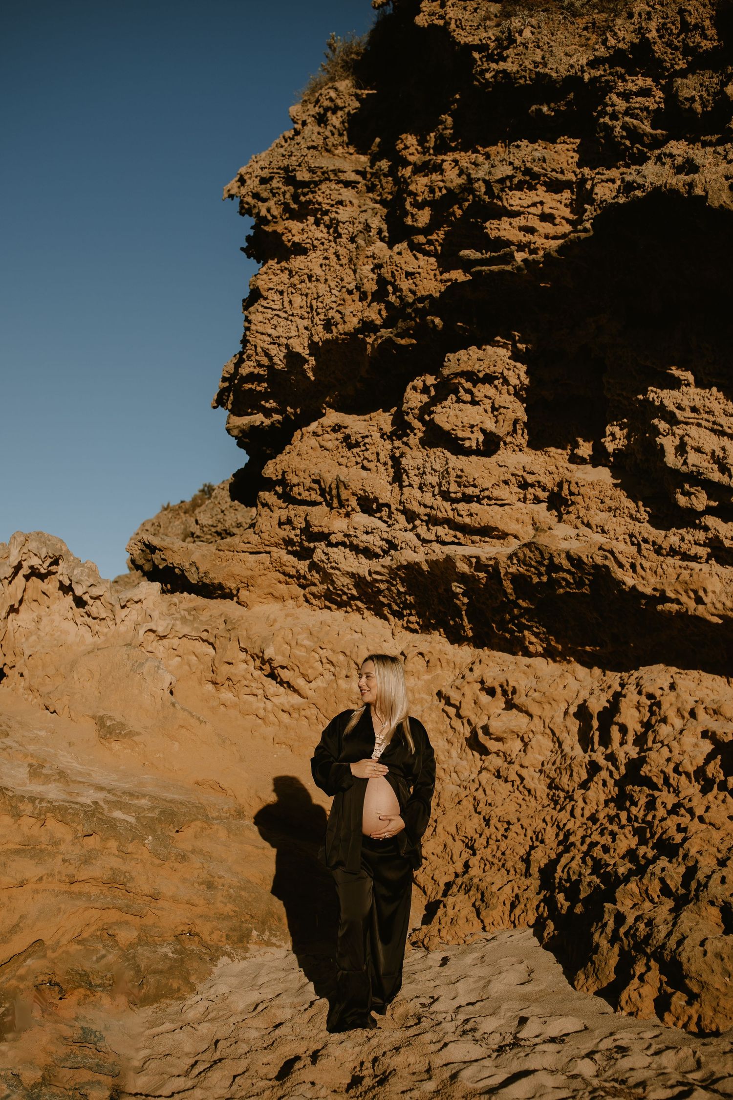 Figure in dark clothing stands against rugged desert cliffs illuminated by warm sunlight.
