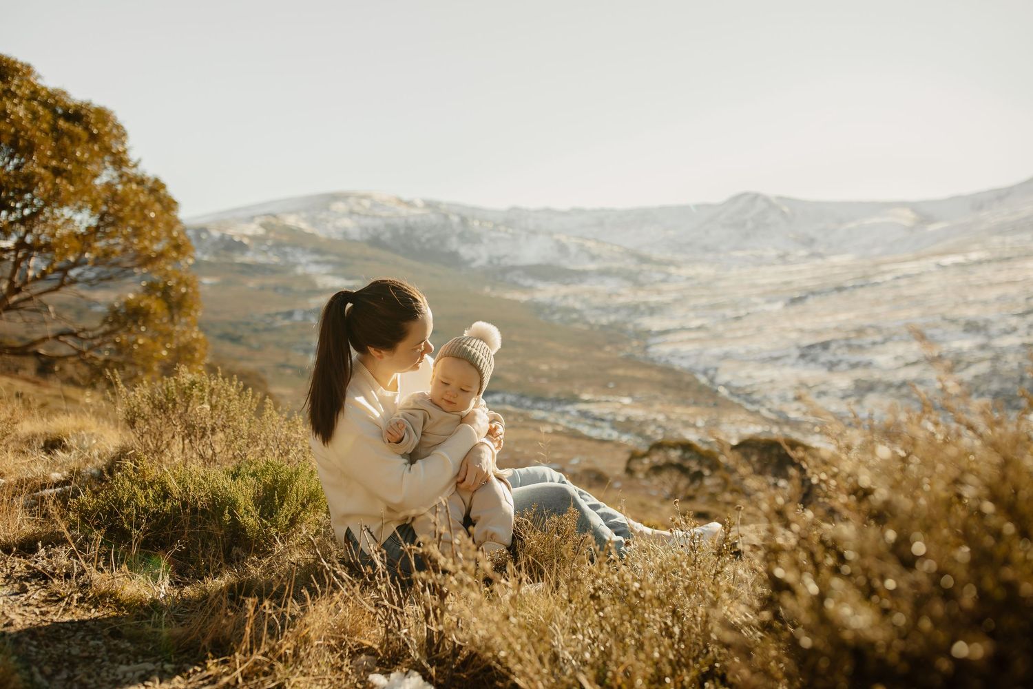 Woman in white sweater sits with young child on grassy hillside overlooking mountainous landscape at sunset.