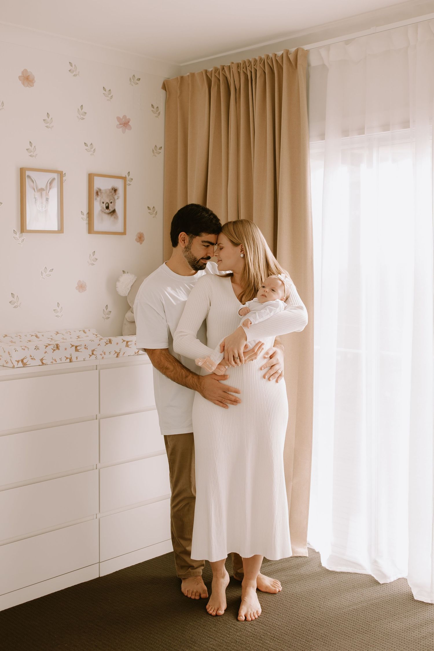 A romantic maternity photoshoot showing a couple embracing in a nursery with floral wallpaper and soft curtains.