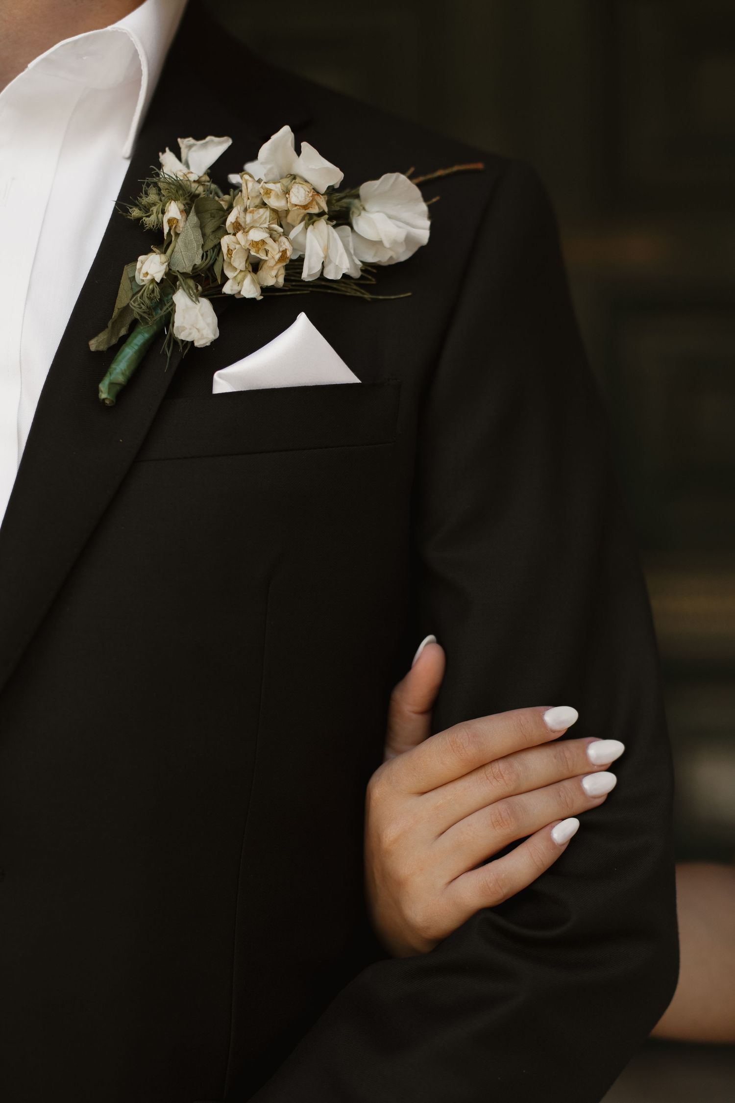 Close-up view of hands with white manicured nails against a black blazer with delicate white floral boutonniere.