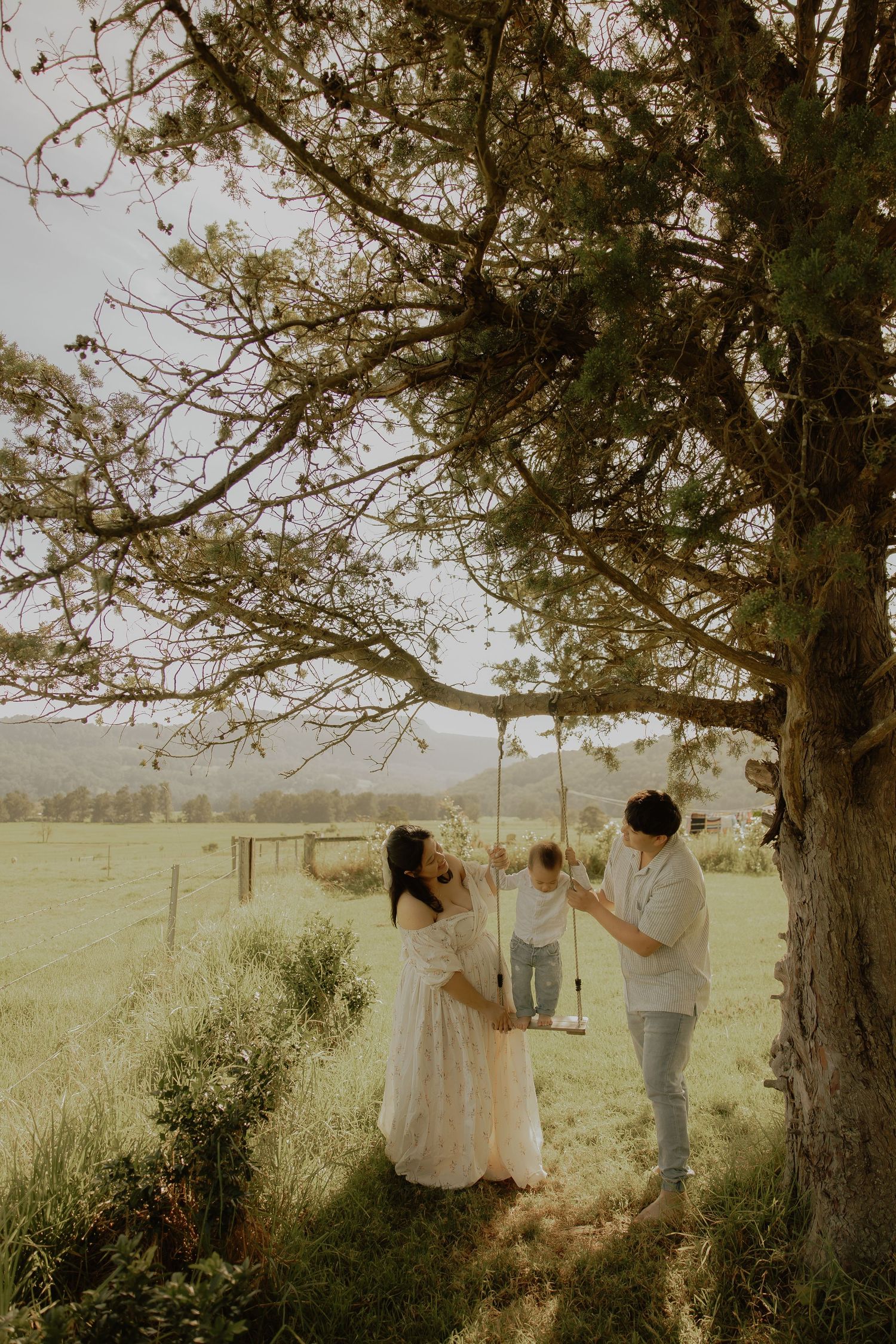 Family with toddler on rope swing under large tree in green pastoral field.