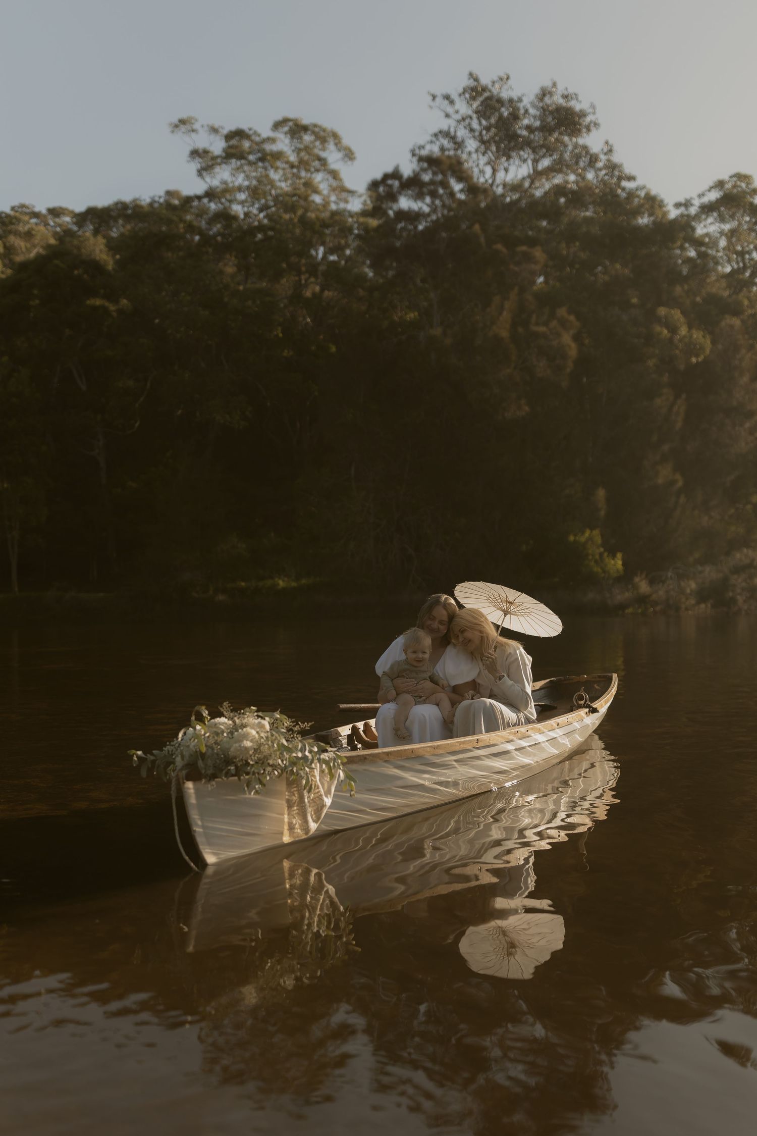 Couple sharing a romantic moment in a white rowboat on a calm lake, holding a parasol, surrounded by lush green trees.