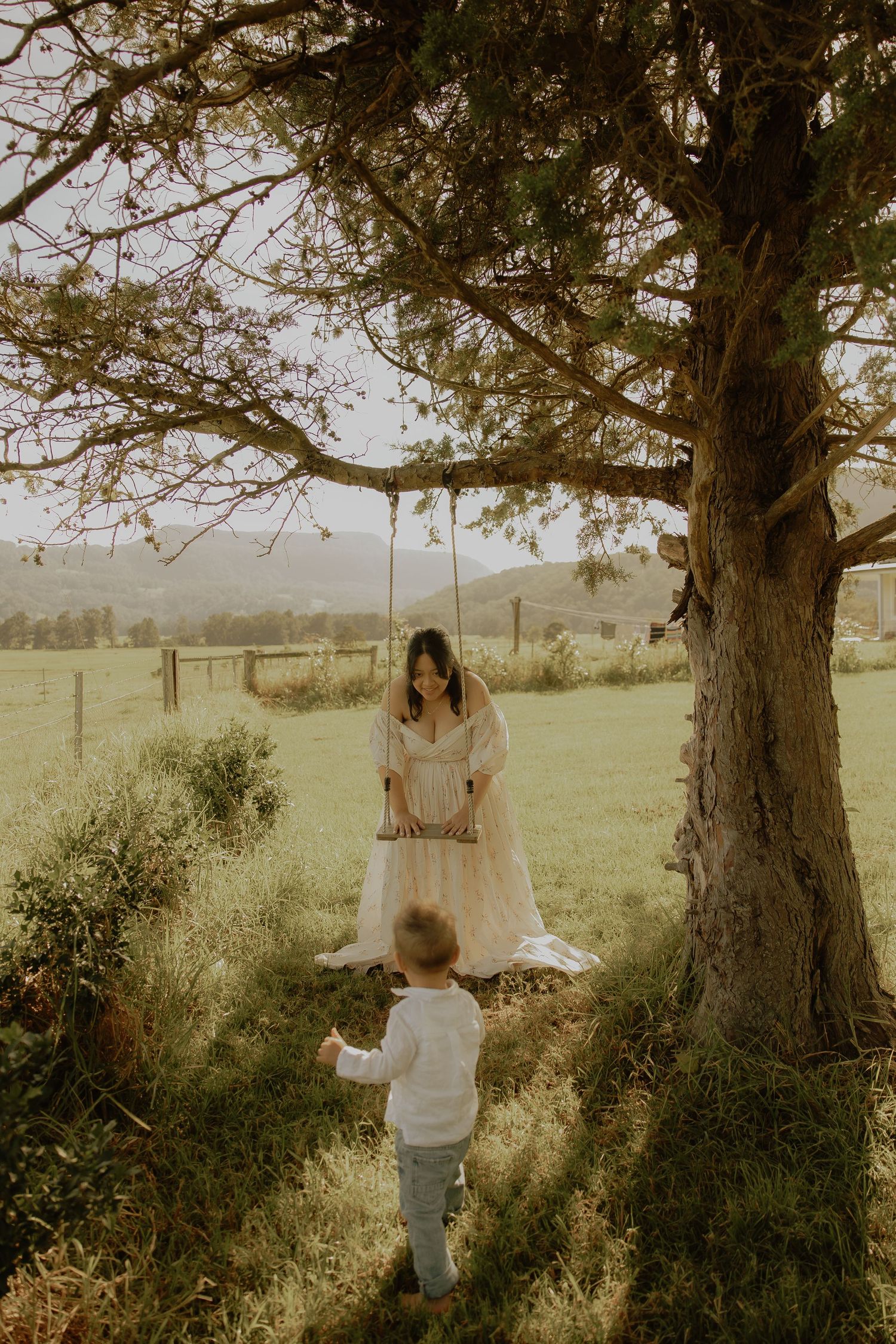 Woman in white gown on a tree swing as a toddler approaches in a green field.