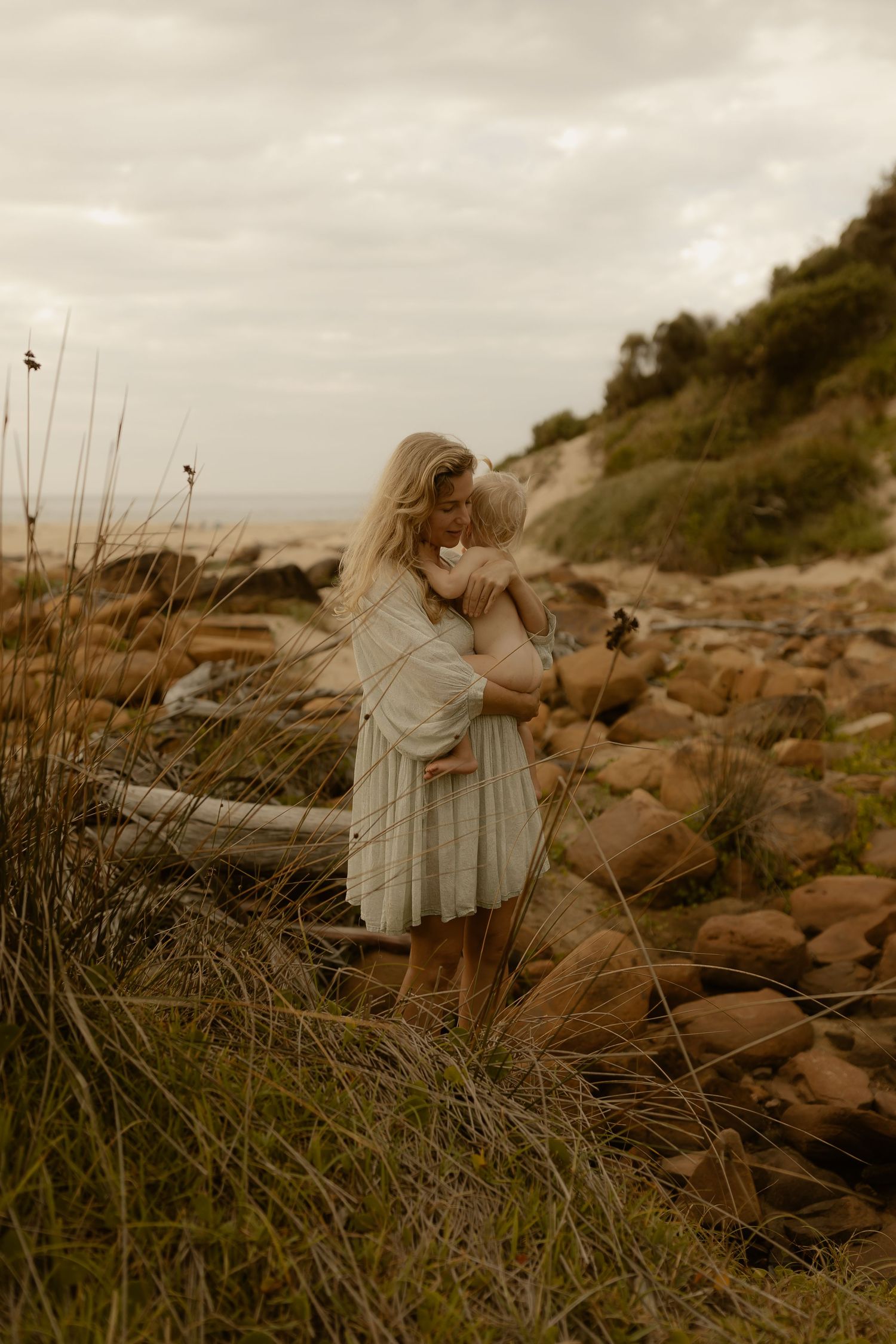 People explore a coastal landscape with tall grass and rocky terrain at sunset.
