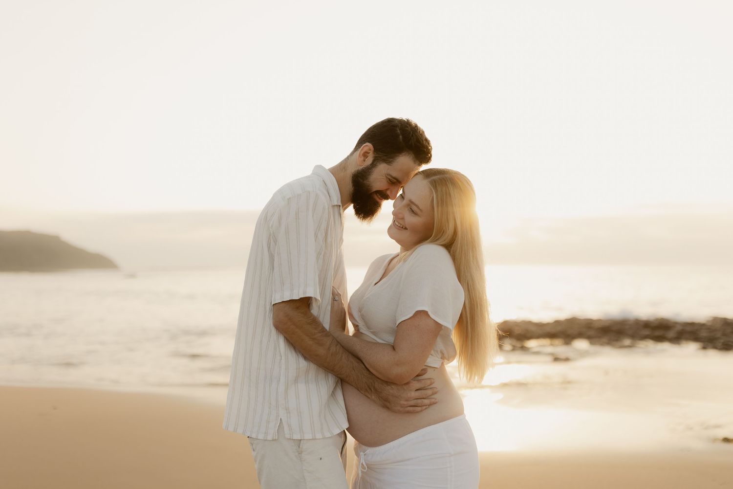 A couple in white outfits embraces tenderly on a beach at sunset with ocean waves in the background.
