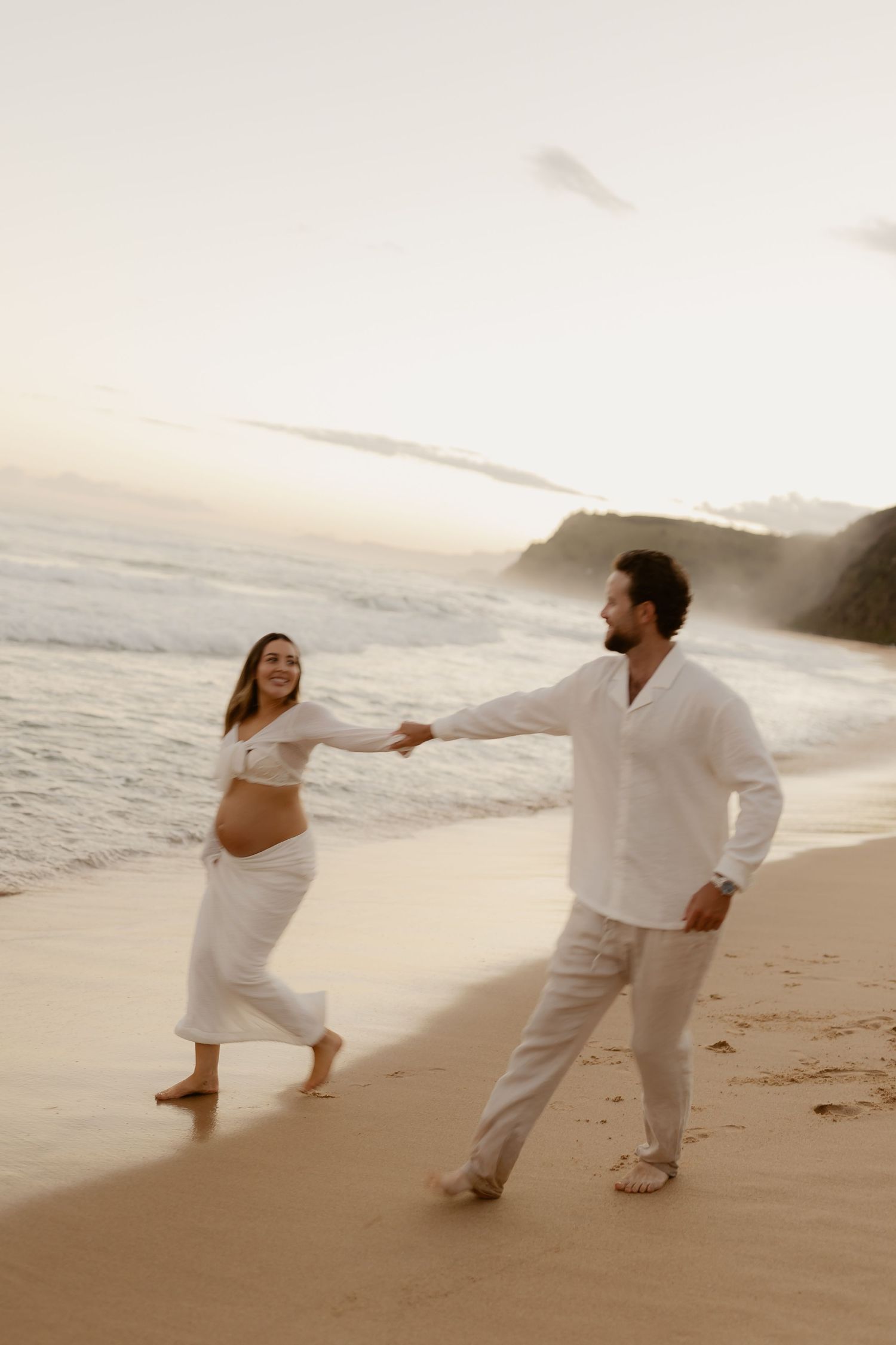 Couple in white clothing playfully dancing together on sandy beach at sunset.