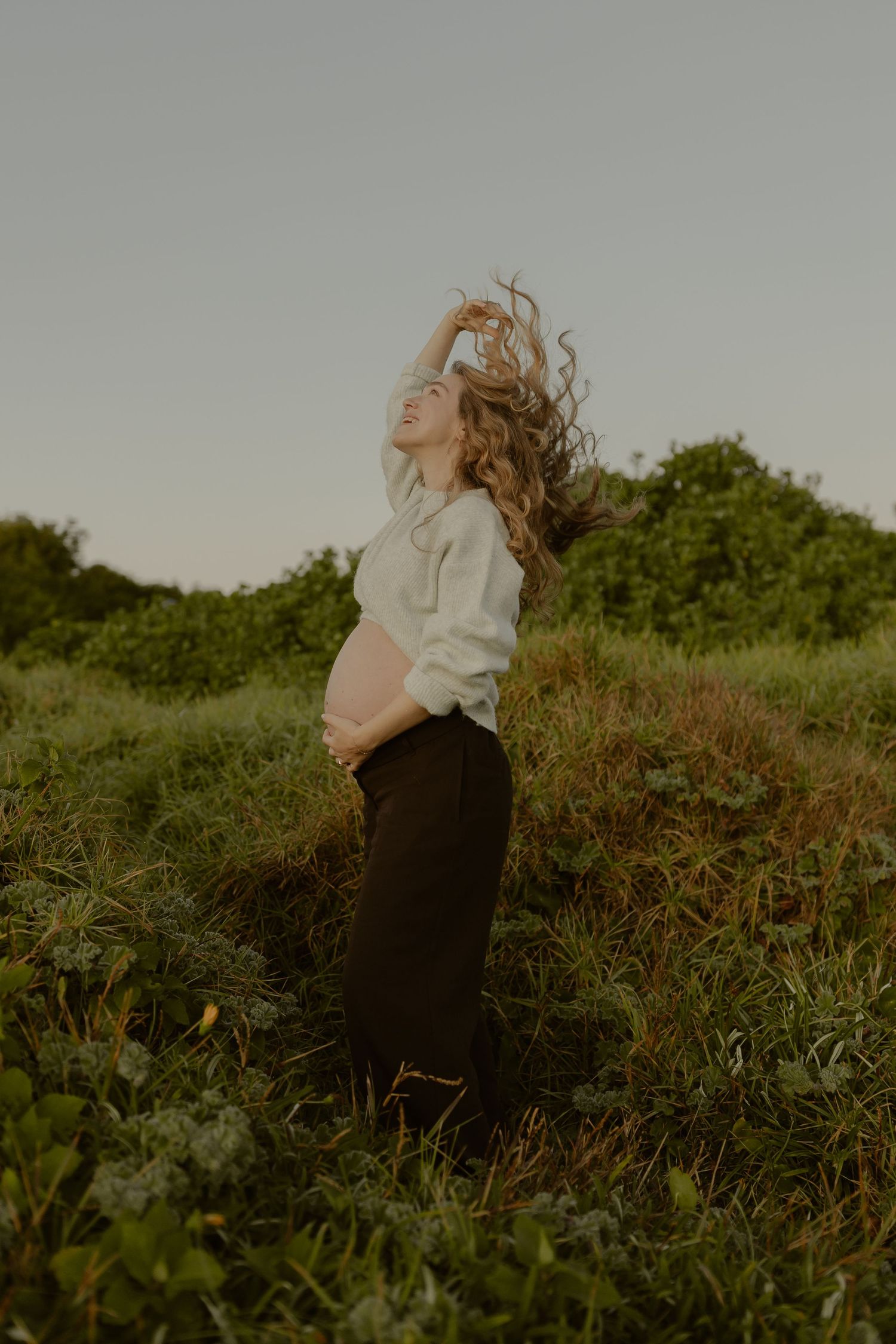 Joyful pregnant woman in white top cradles baby bump in lush green field at sunset.