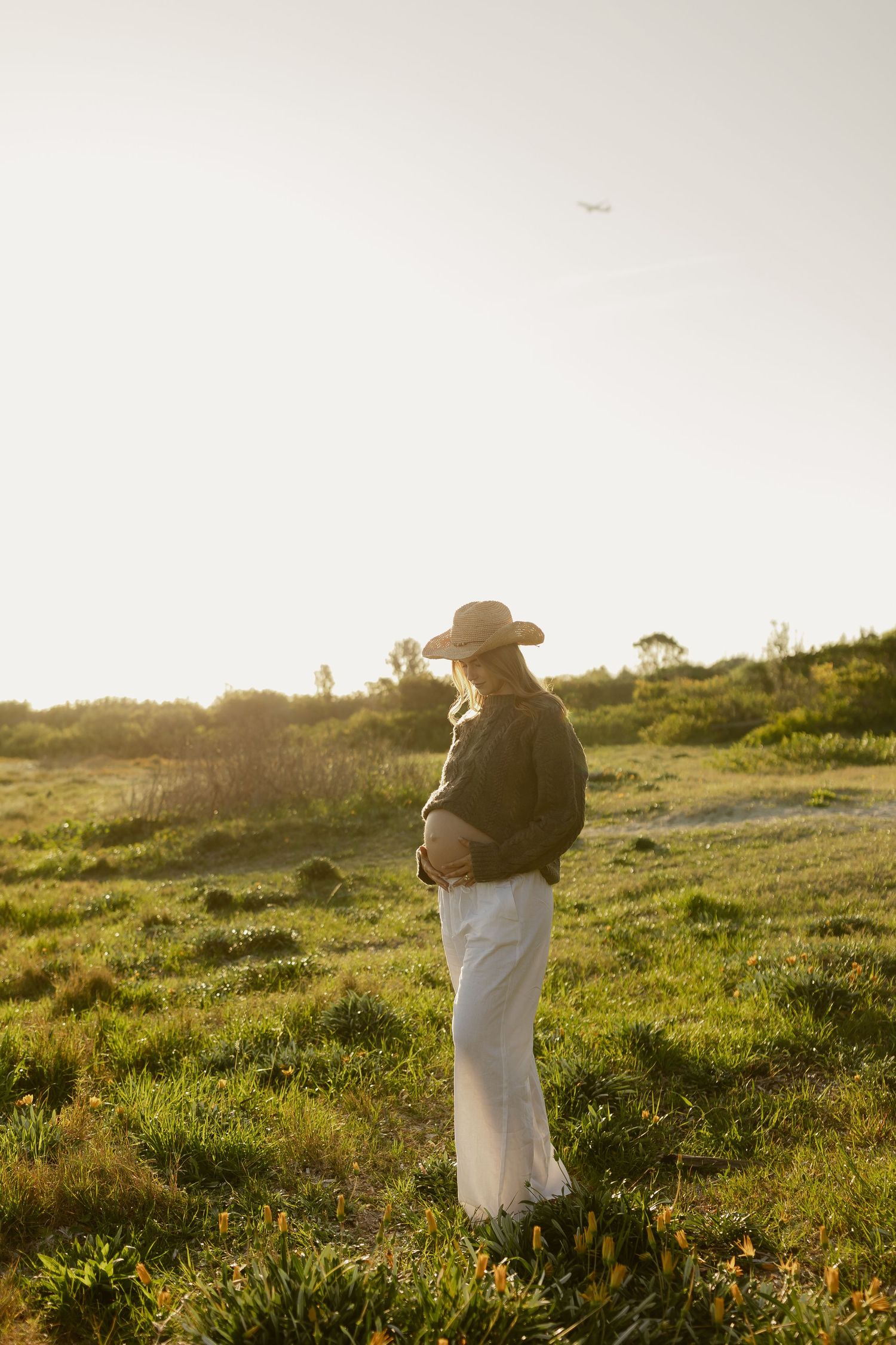 Pregnant woman in white pants, green jacket, and straw hat standing in a sunlit meadow, cradling her baby bump.