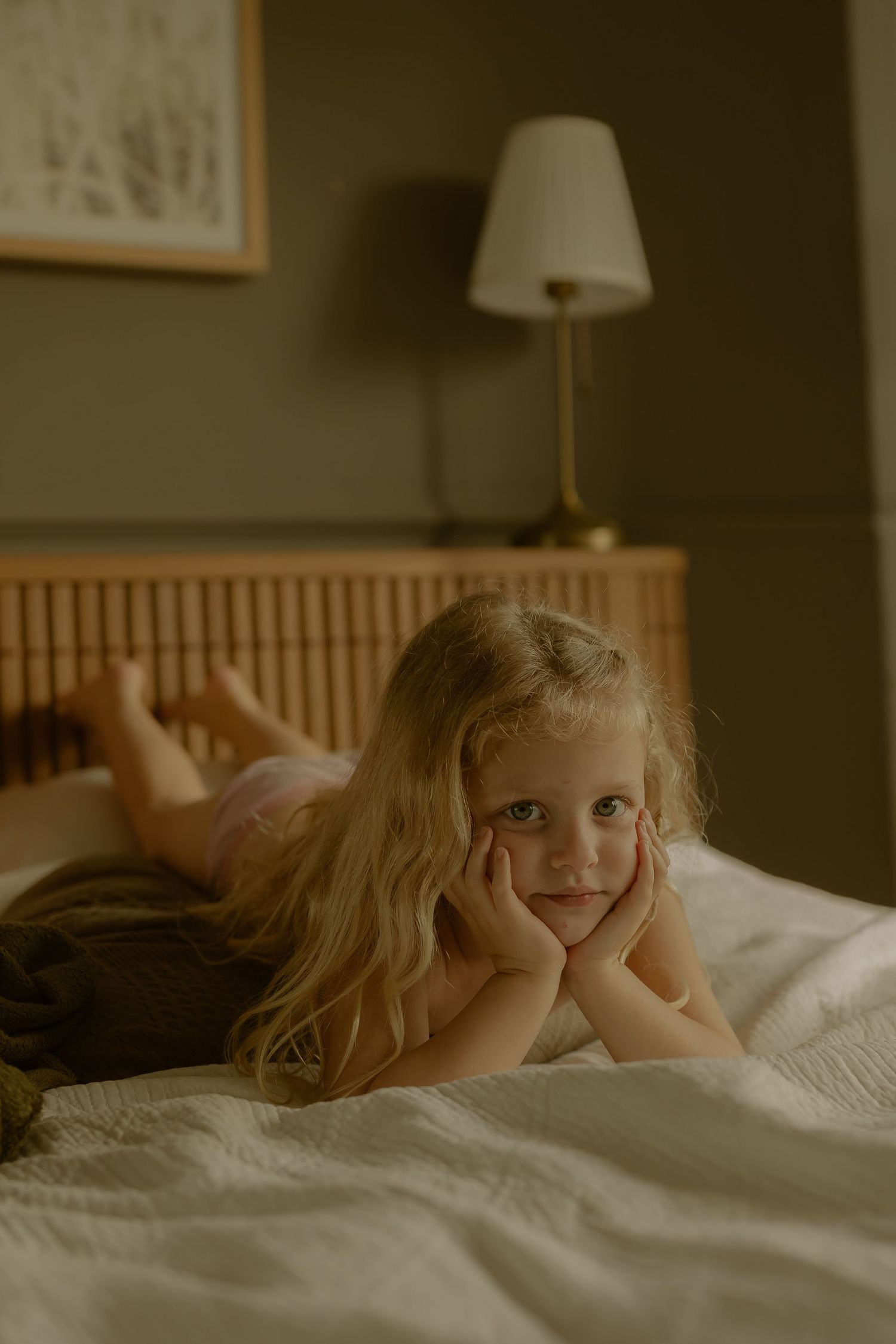 Blonde little girl lying on a white bed with hands on cheeks, looking at camera in a cozy bedroom.