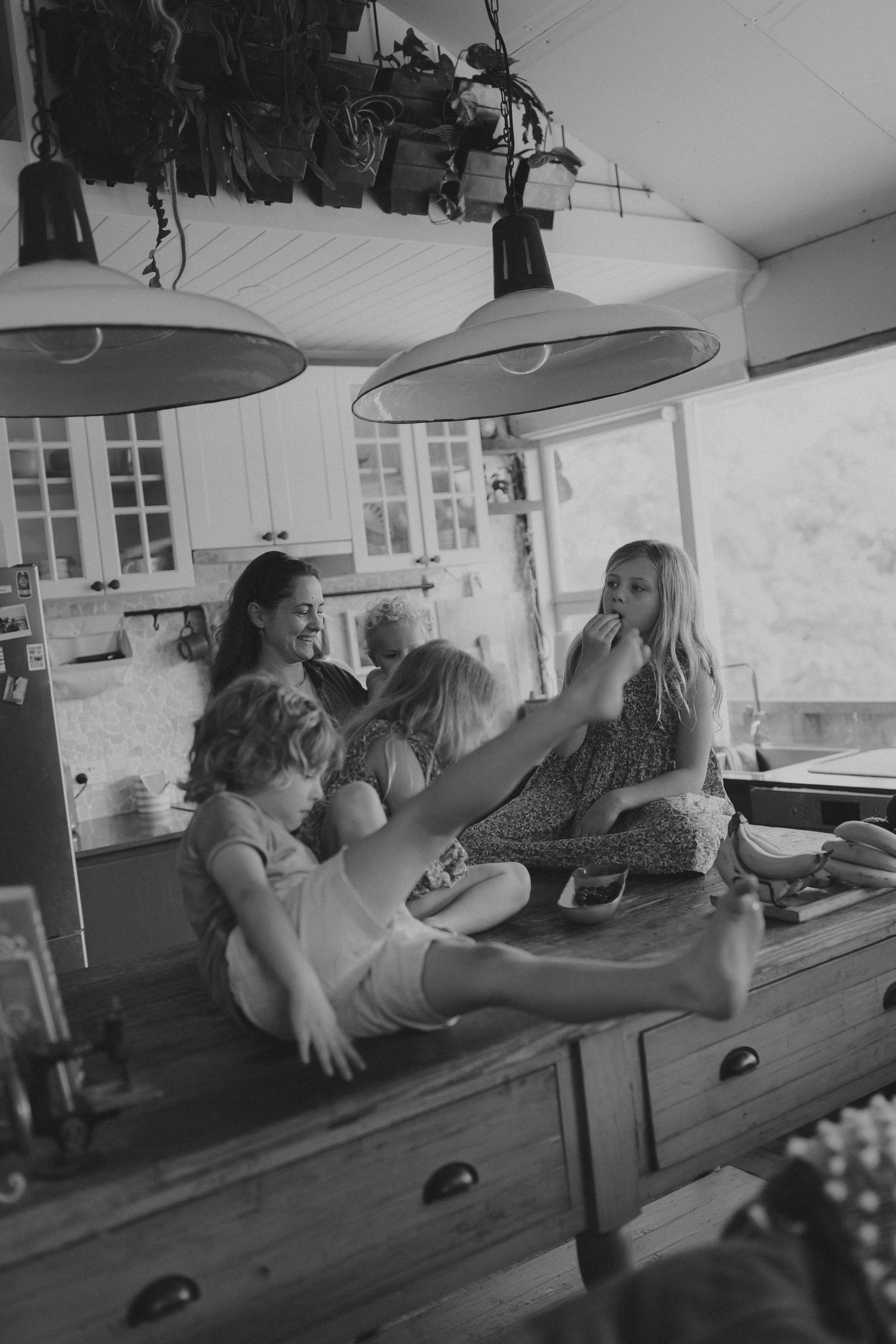 Black and white photo of a mother and children sitting on a wooden kitchen counter in a cozy farmhouse kitchen.