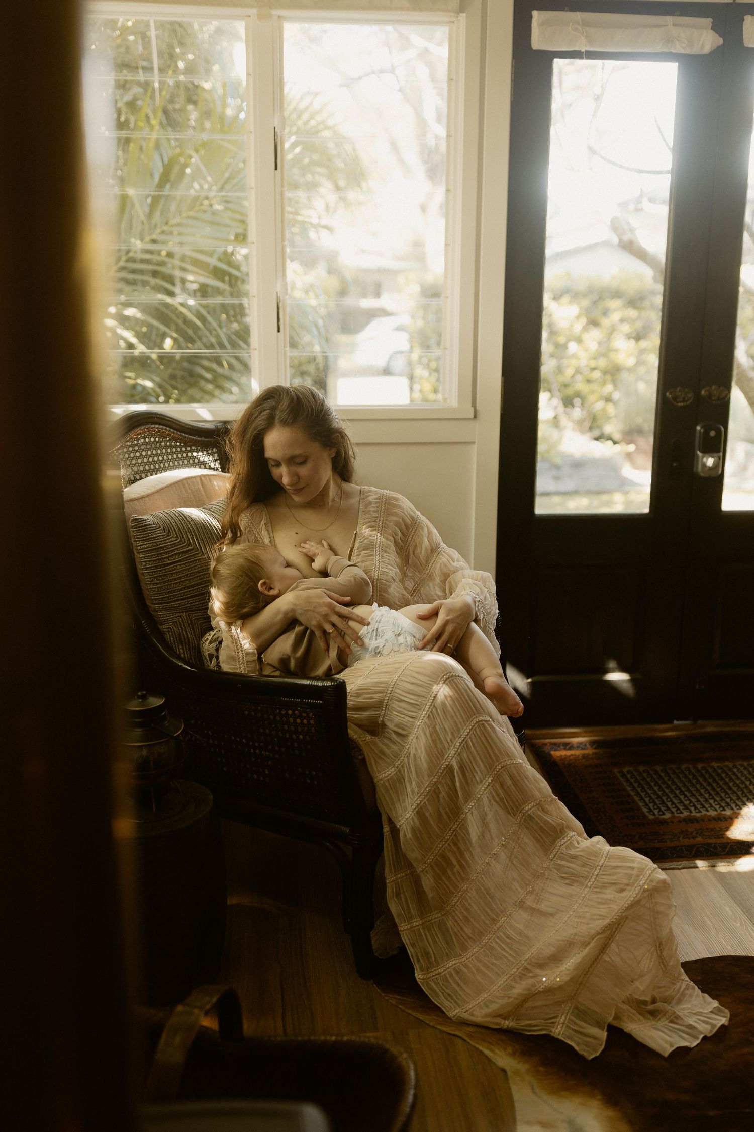 Person in flowing white dress resting on wicker chair in sunlit room.