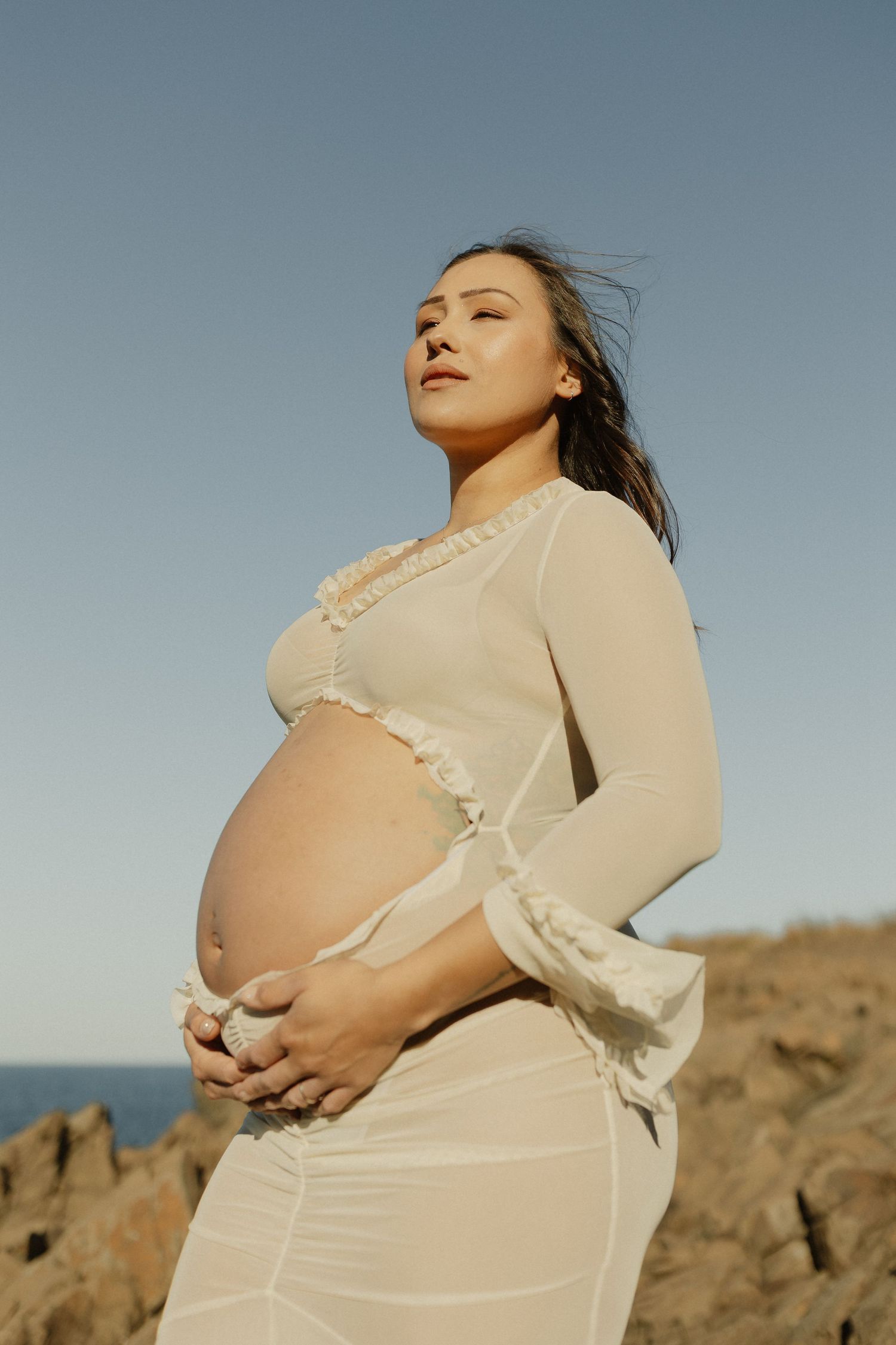 Maternity portrait of a woman in a sheer white dress posing against a coastal backdrop during golden hour.