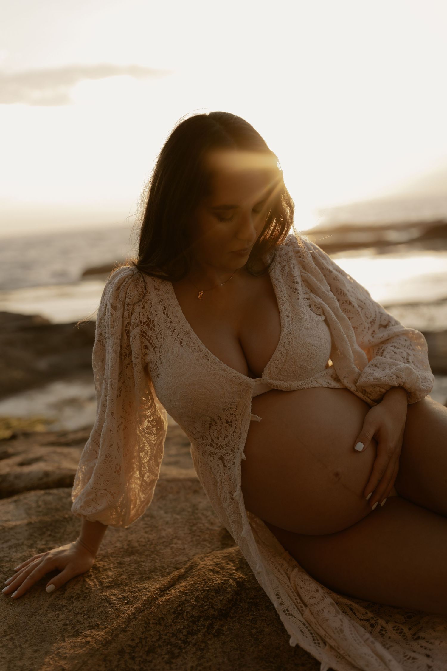 Maternity photoshoot at sunset on beach with woman in white lace dress reclining on sand in warm golden light.