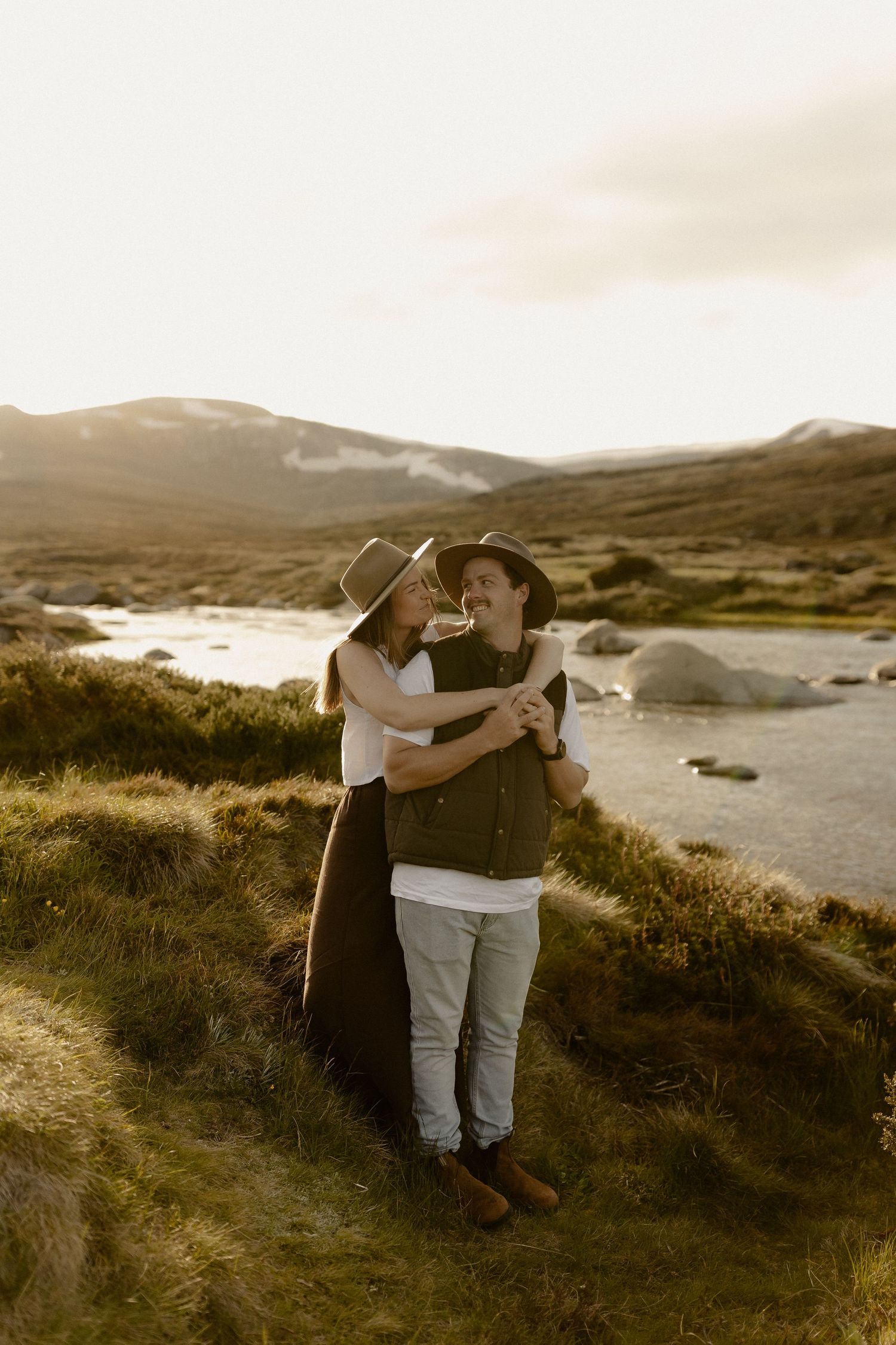 A romantic couple embraces on a grassy hillside overlooking a scenic lake during golden hour in a mountainous landscape.