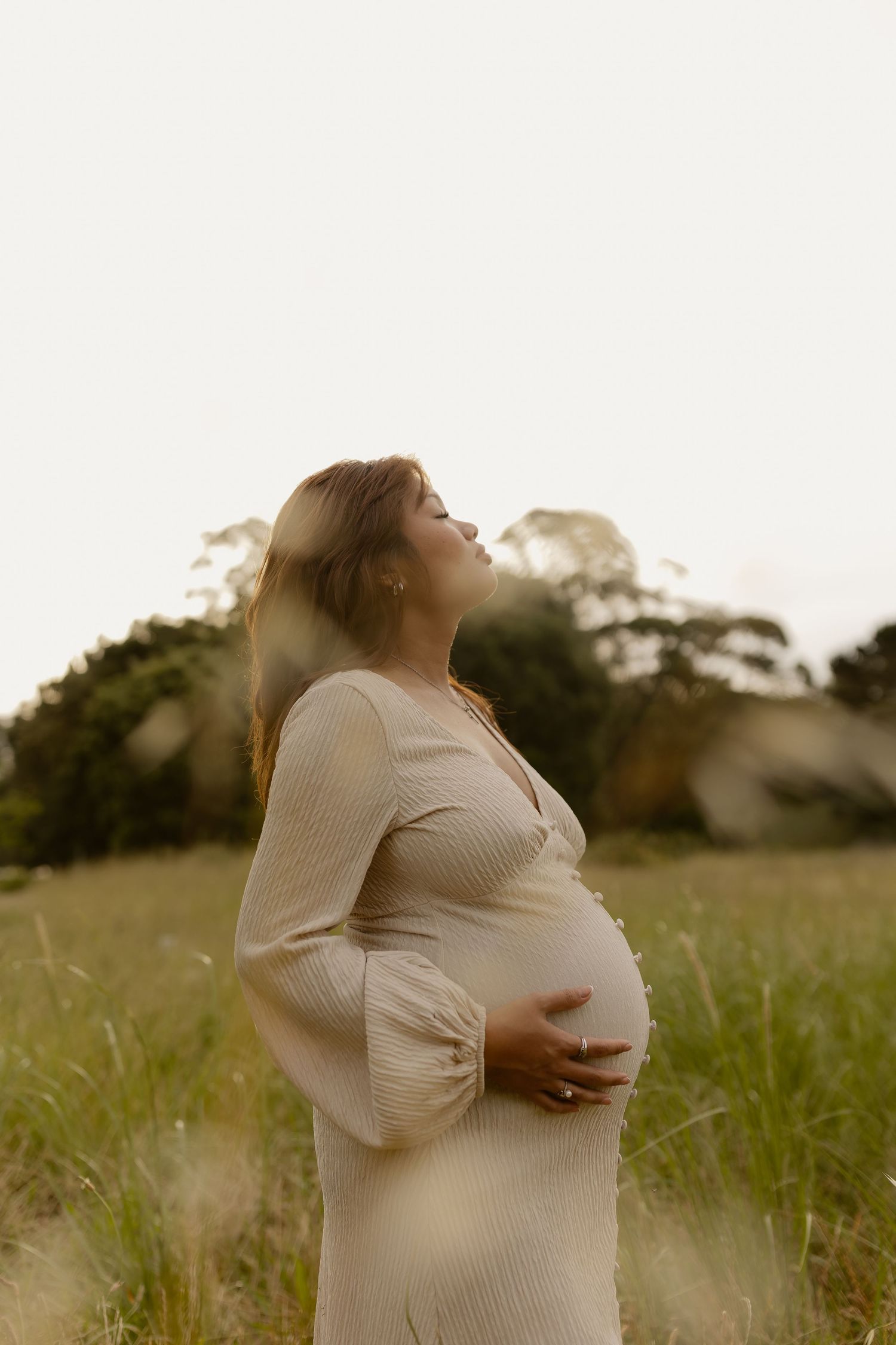 A pregnant woman in a white dress stands in a sunlit meadow during golden hour.