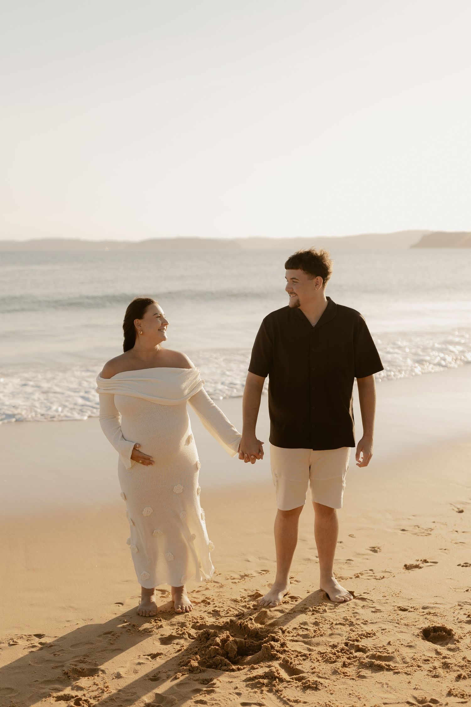 A couple walking hand in hand on a sandy beach during golden hour.