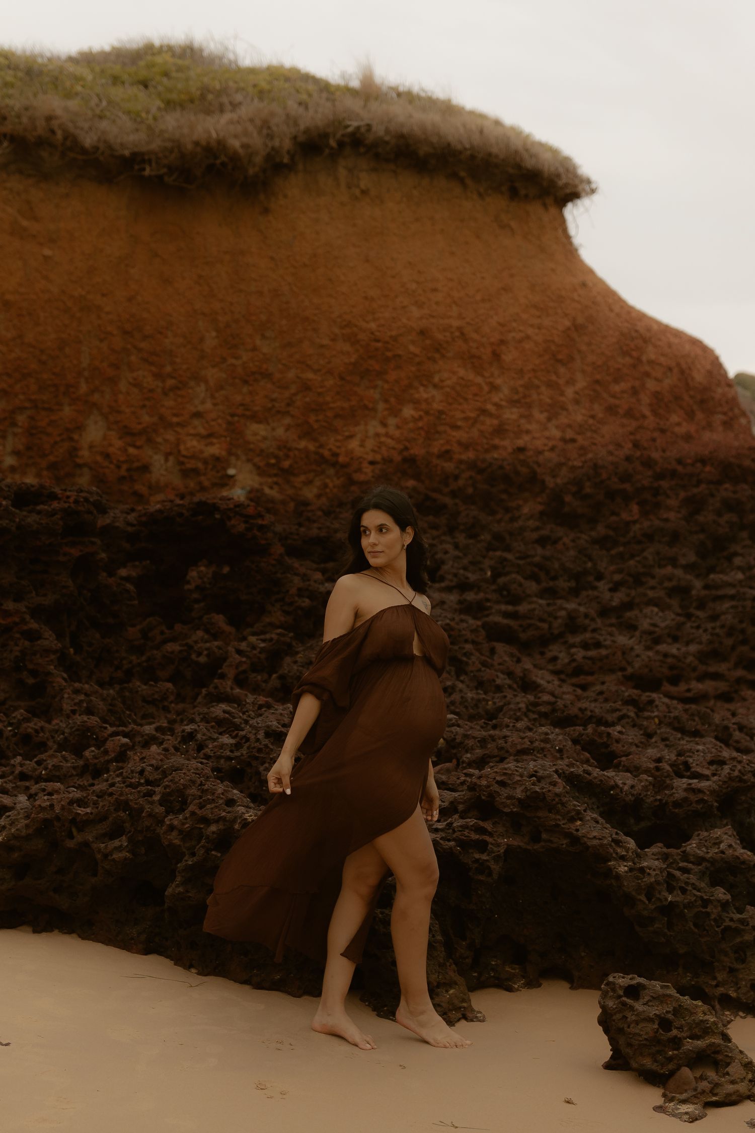 Pregnant woman in brown dress poses near dramatic coastal cliffs during golden hour photography session.