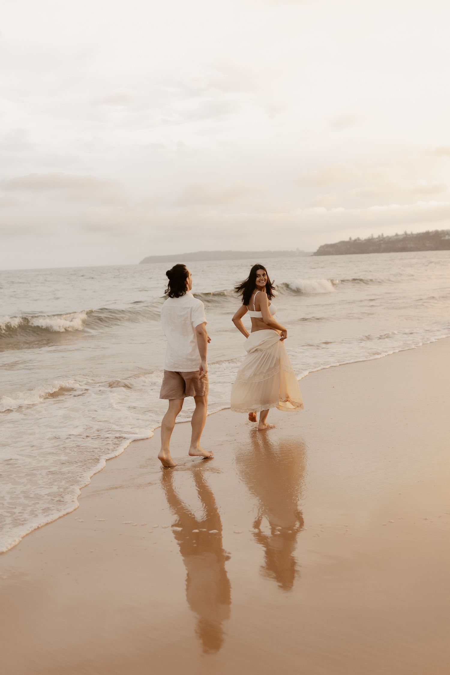Couple running playfully on sandy beach at sunset with reflections in wet sand.