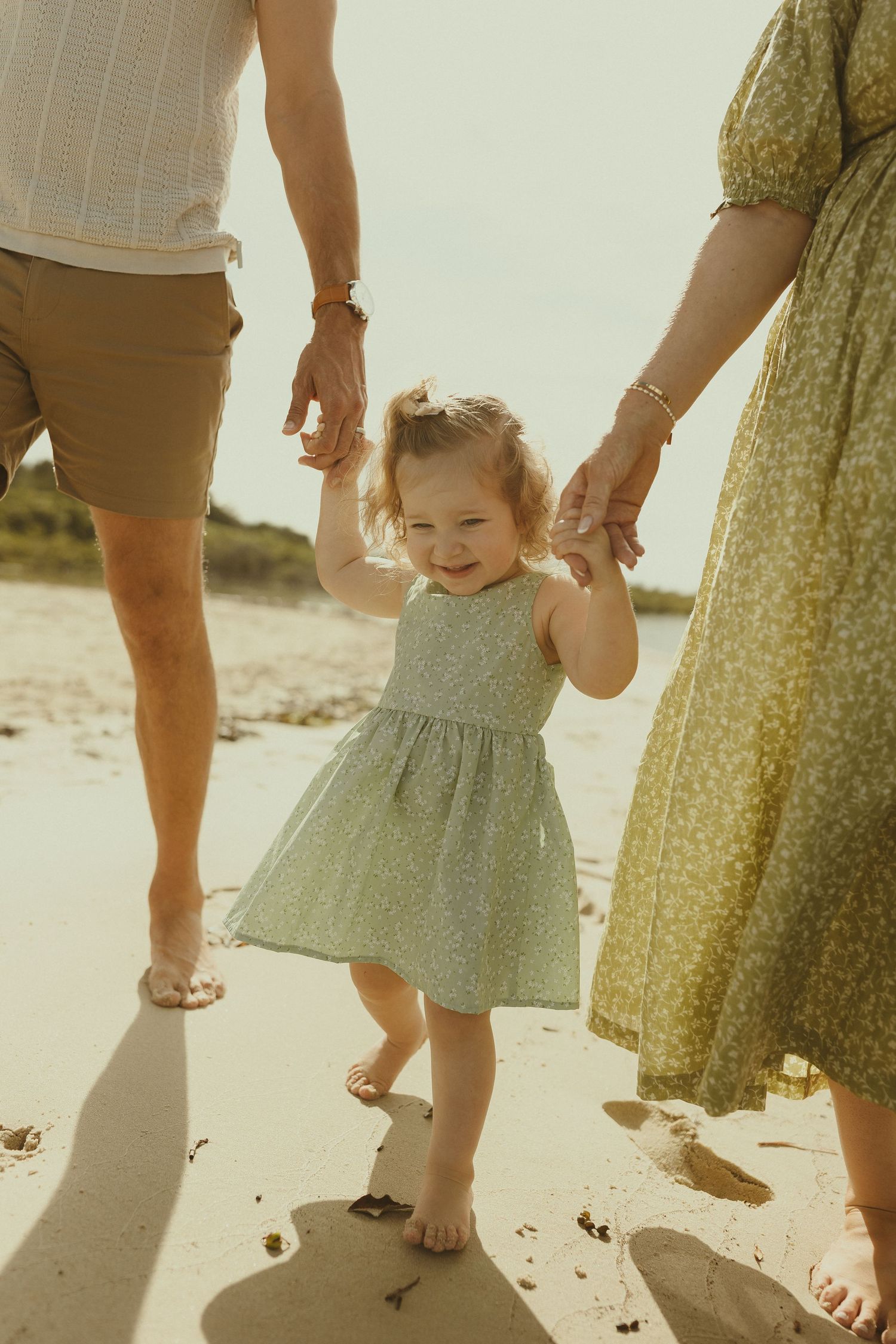 A toddler walks along the beach holding hands with parents, their feet in the sand.