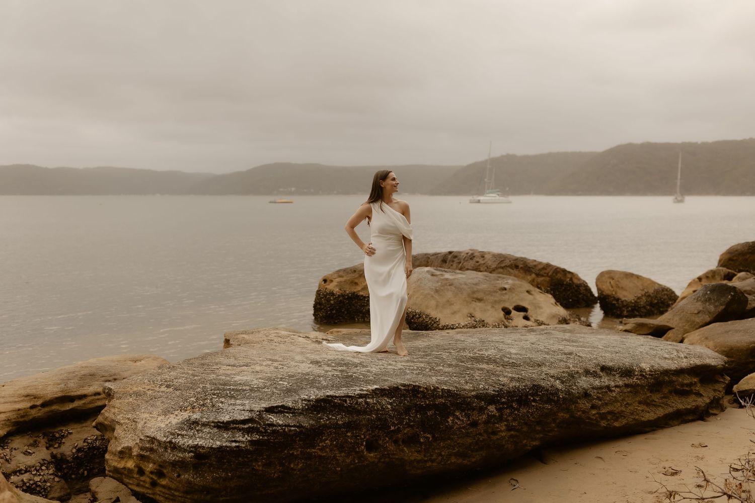 A person in white stands on a rocky coastal overlook with mountains and water in the distance.