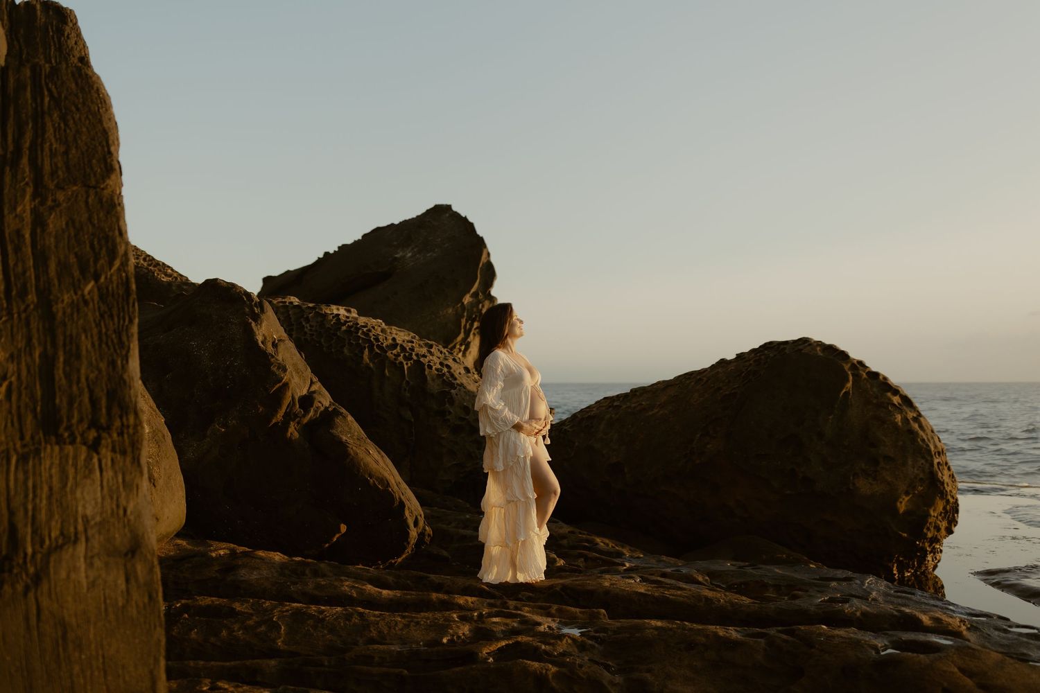 A figure in a flowing white dress stands on coastal rocks at sunset against a misty ocean backdrop.