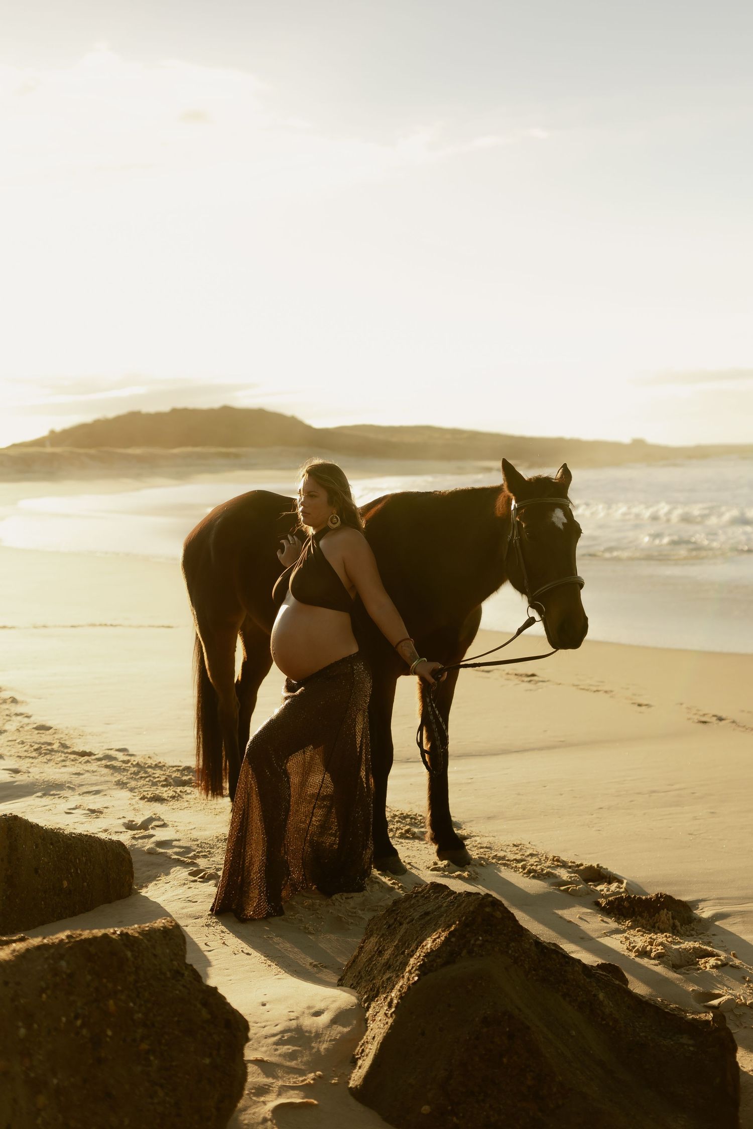 Pregnant woman in sheer gown stands with dark horse on sandy beach at golden sunset.