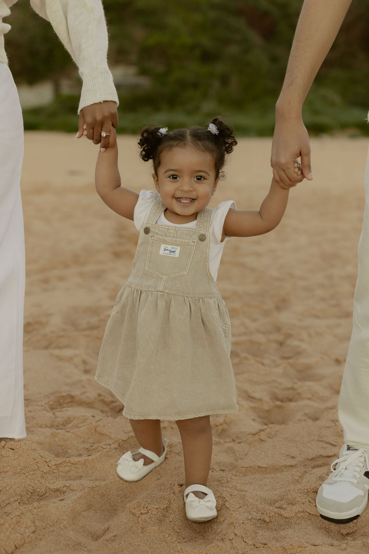 Smiling toddler girl in linen pinafore dress holding parents hands on sandy beach.