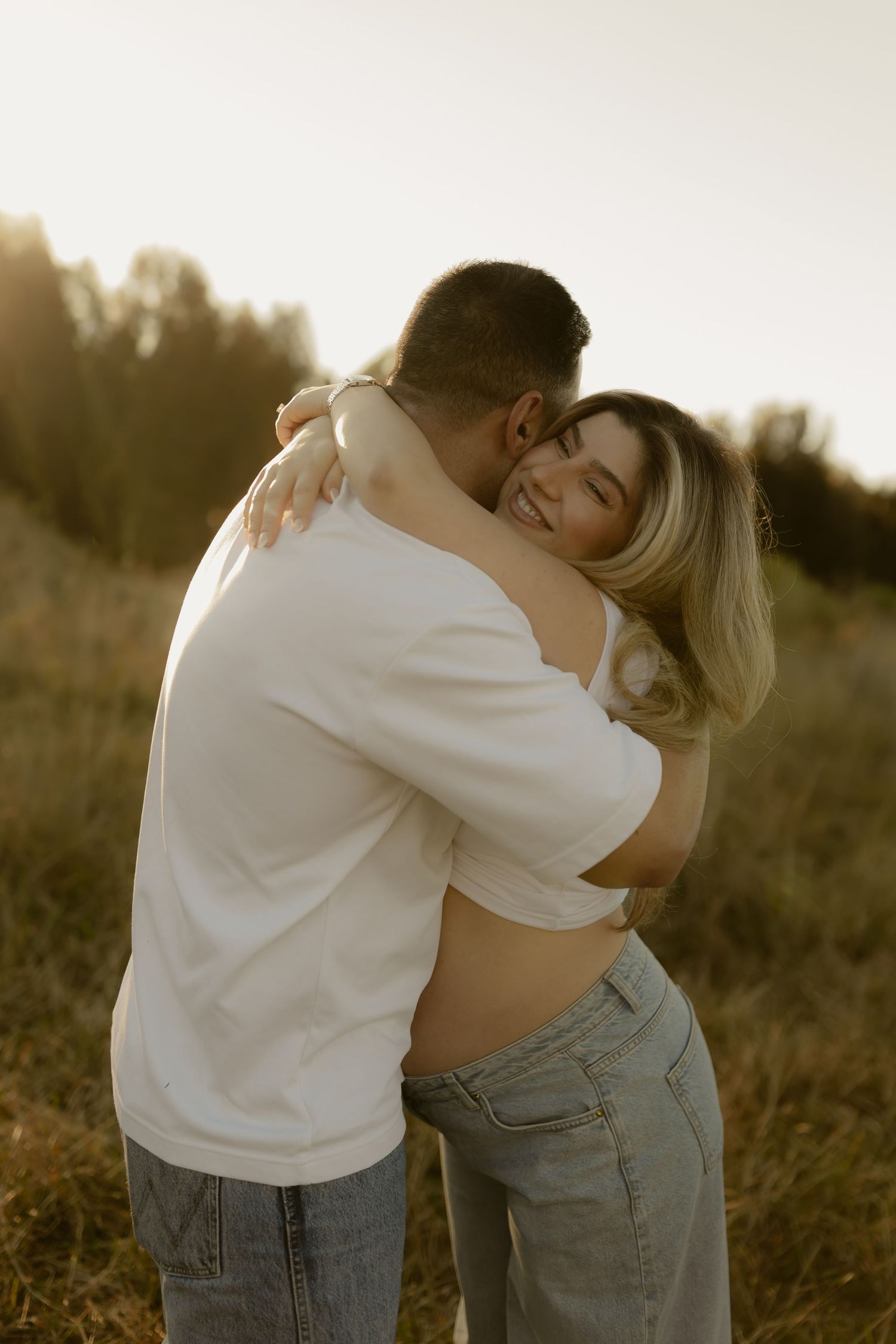 Smiling pregnant woman embracing man in white t-shirt in sunlit field during golden hour maternity shoot.