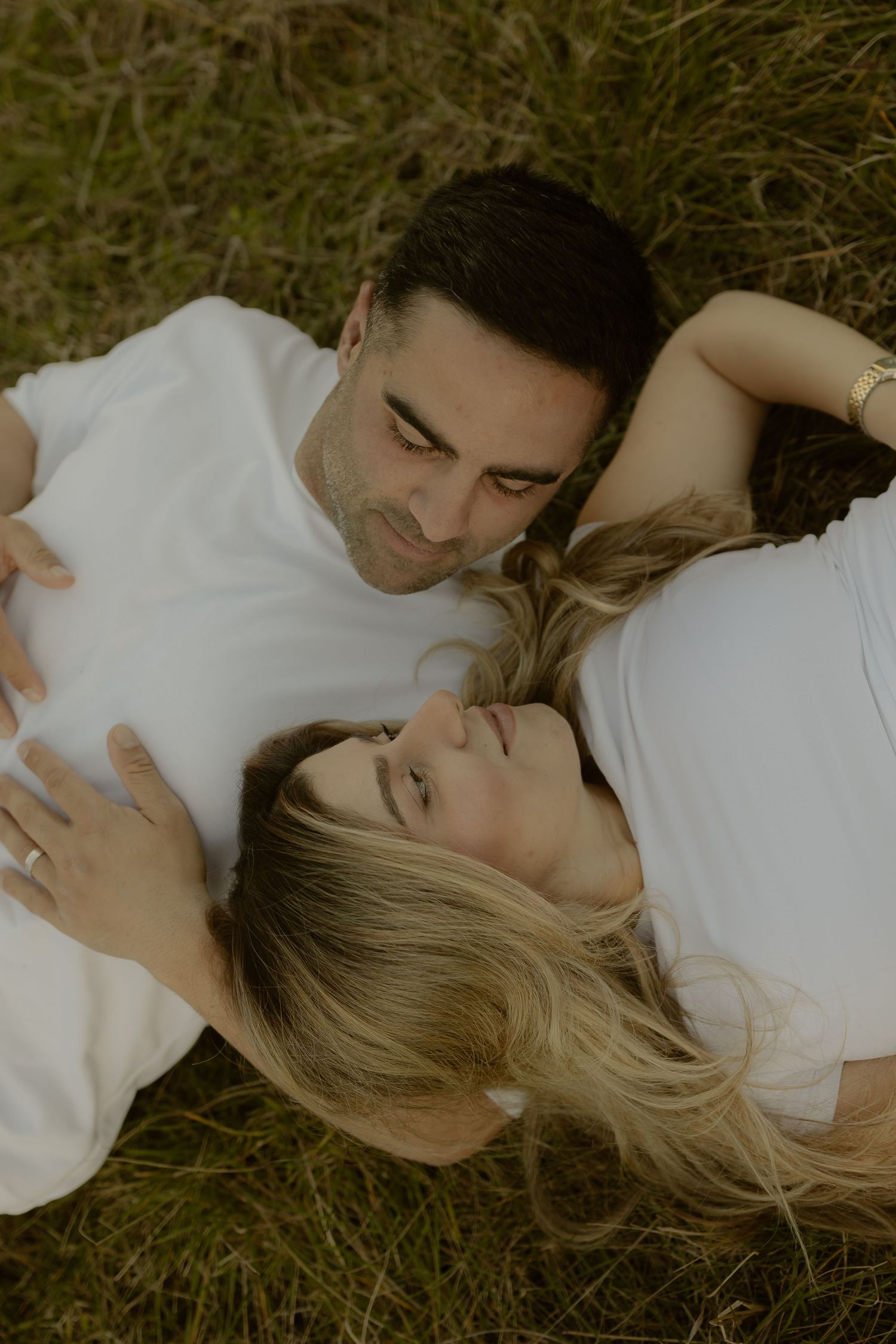 Aerial view of couple lying in green grass wearing white shirts, man resting head on pregnant belly.