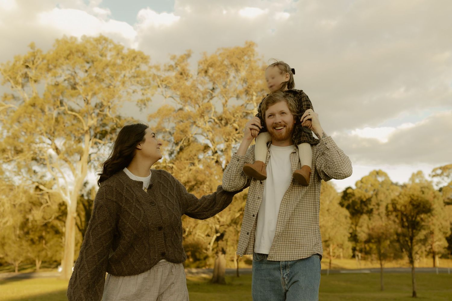 A happy family enjoying a park walk, with a bearded dad carrying a child on his shoulders beside a smiling woman.