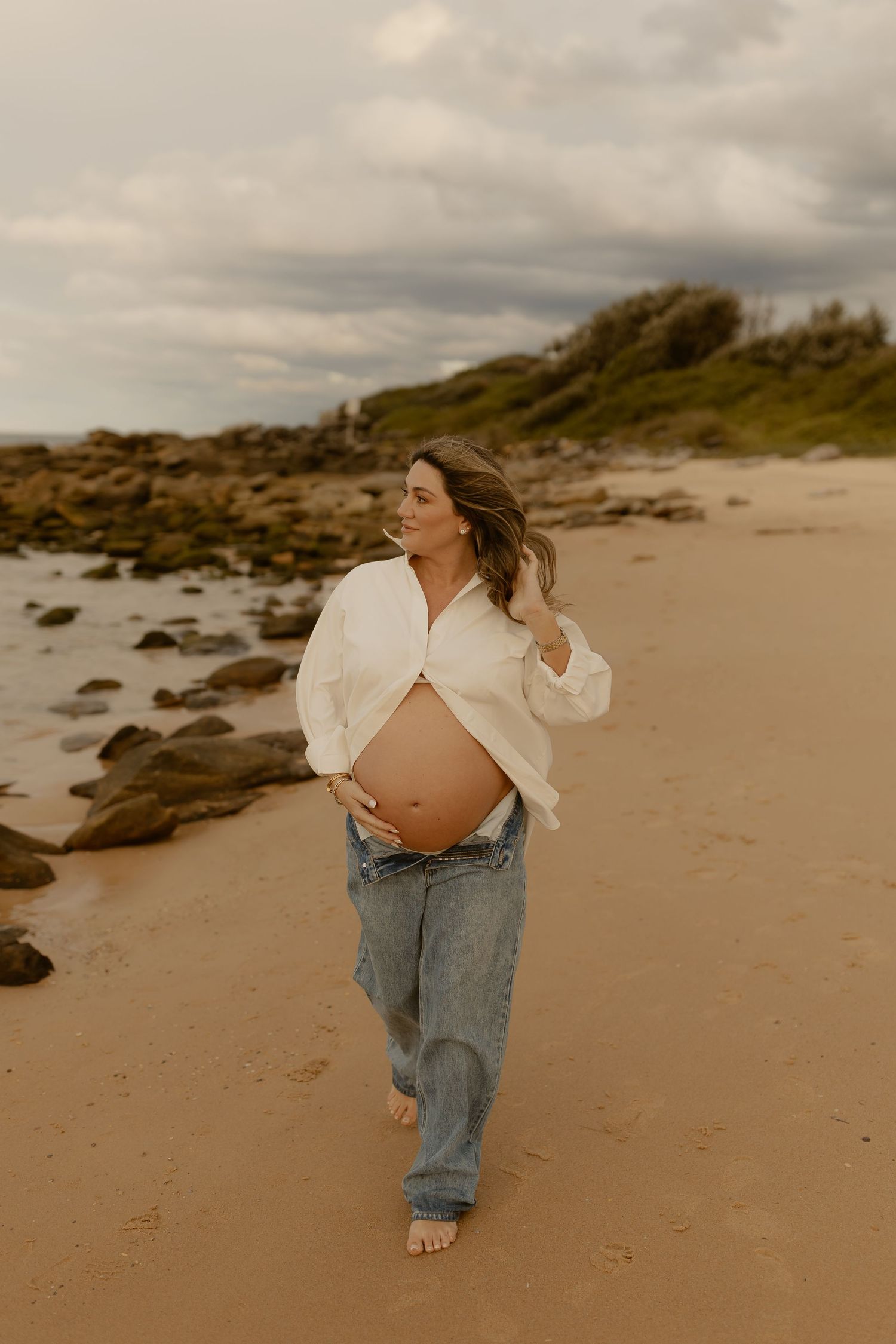 Pregnant woman in white shirt and jeans cradling baby bump on sandy beach with rocky coastline and cloudy sky.
