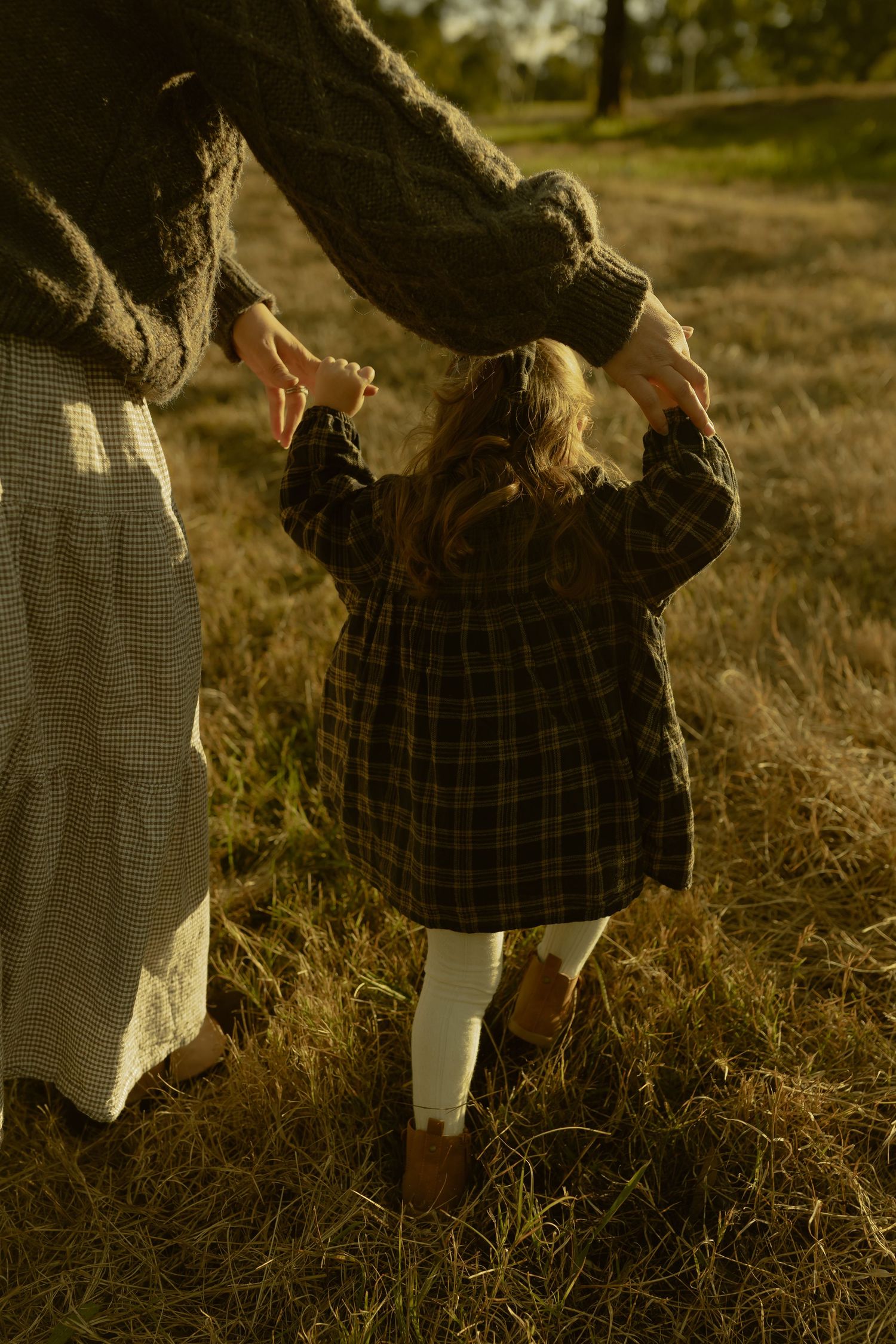 A young girl in a plaid dress holds her mother's hands while walking through a golden sunlit field in autumn.