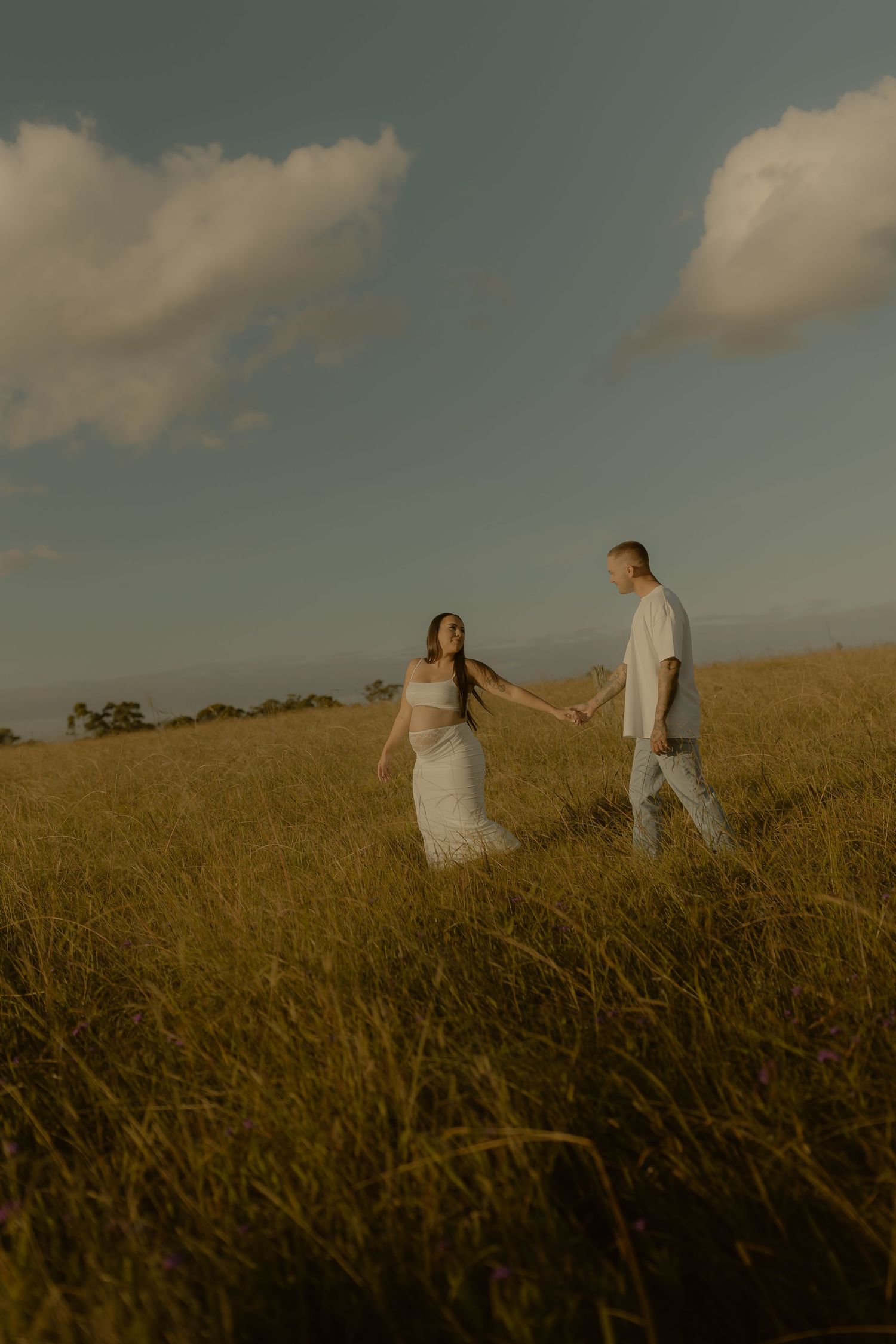 A couple holds hands walking through a golden grass field under a blue sky with clouds at sunset.