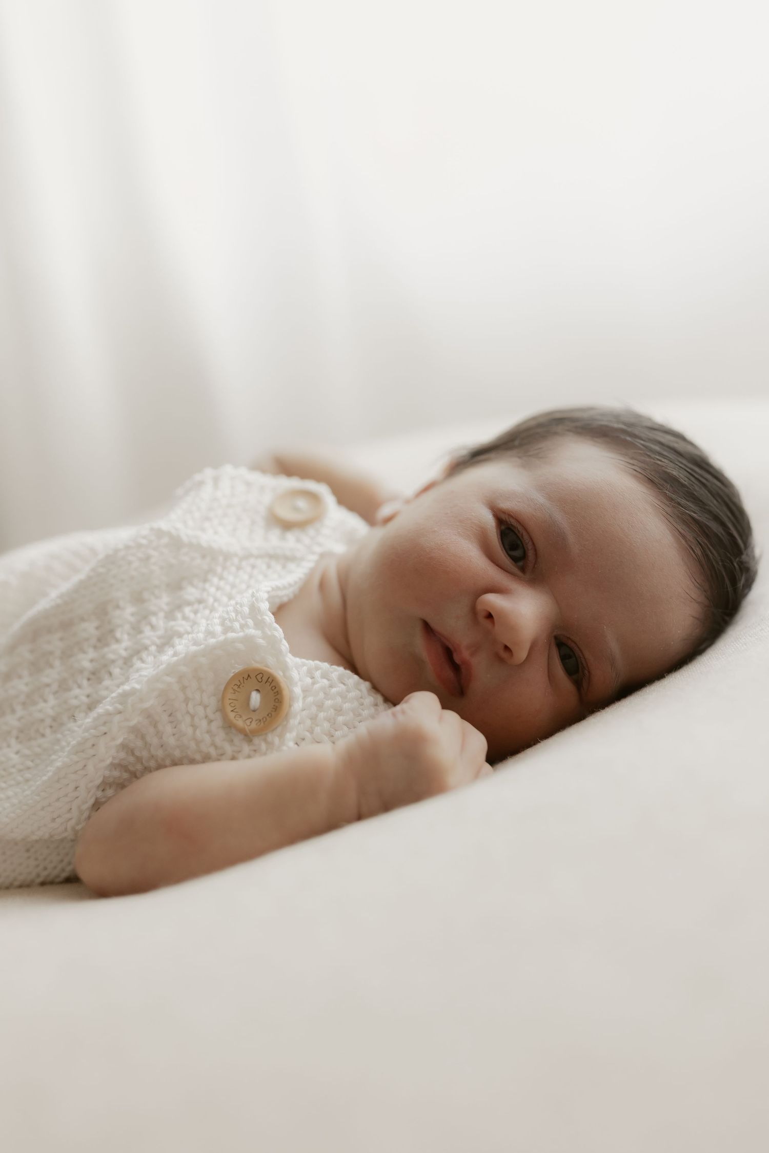 A newborn baby in a white knit romper with wooden buttons lies on a soft white surface, gazing gently at the camera.