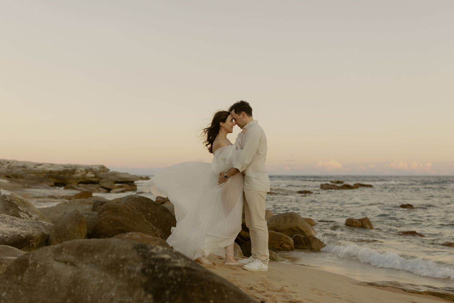 Couple embracing on rocky beach at sunset, woman in flowing white gown near ocean waves.