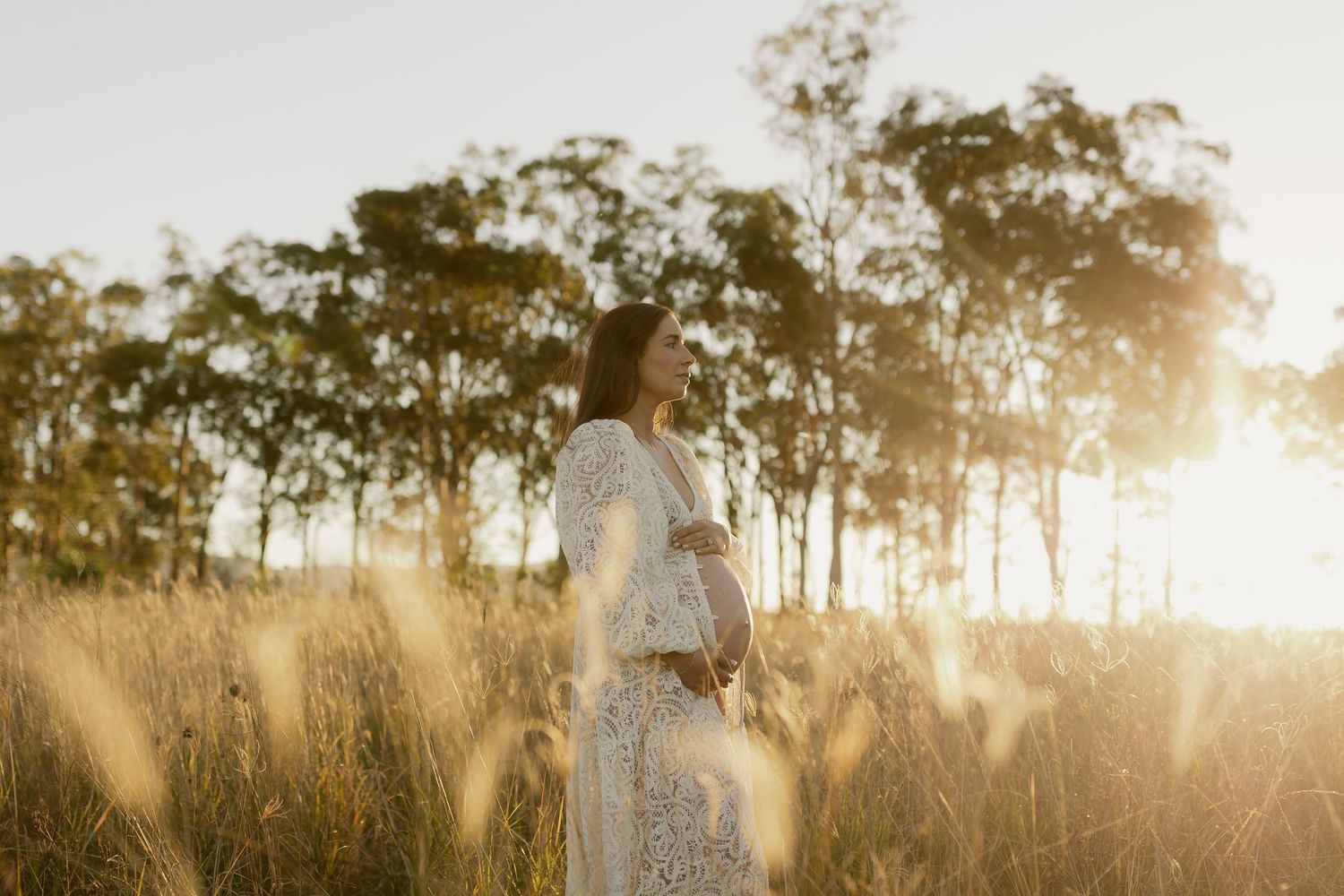 Pregnant woman in lace dress standing in sunlit field with trees at golden hour.