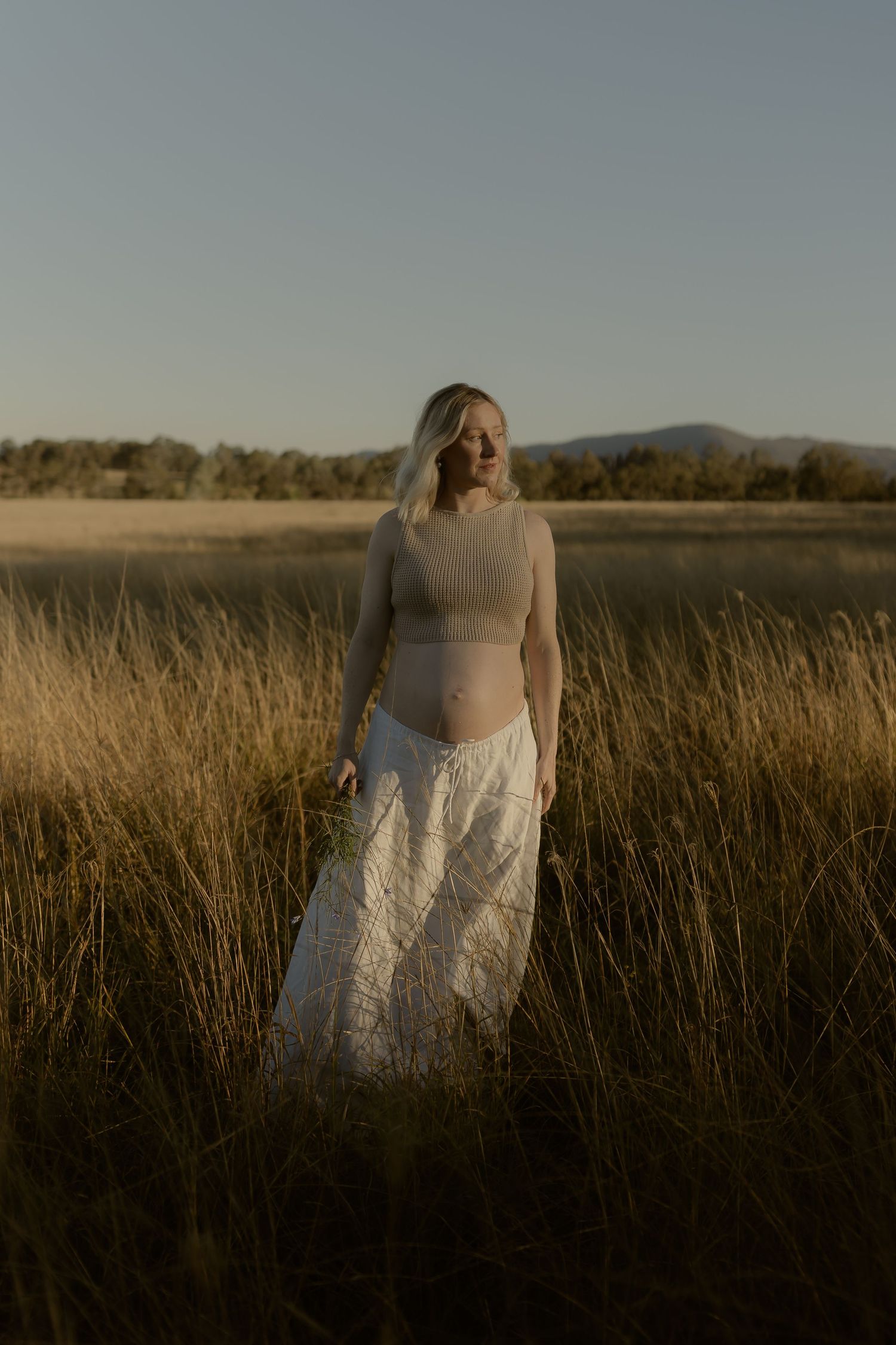 Pregnant woman in crop top and white lace skirt standing in golden grass field at sunset with mountains in background.
