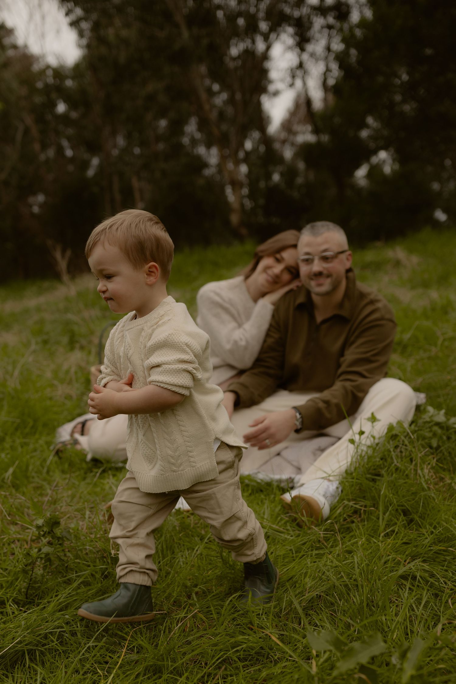 Toddler walks independently on green grass while parents sit relaxed together on a blanket behind him.