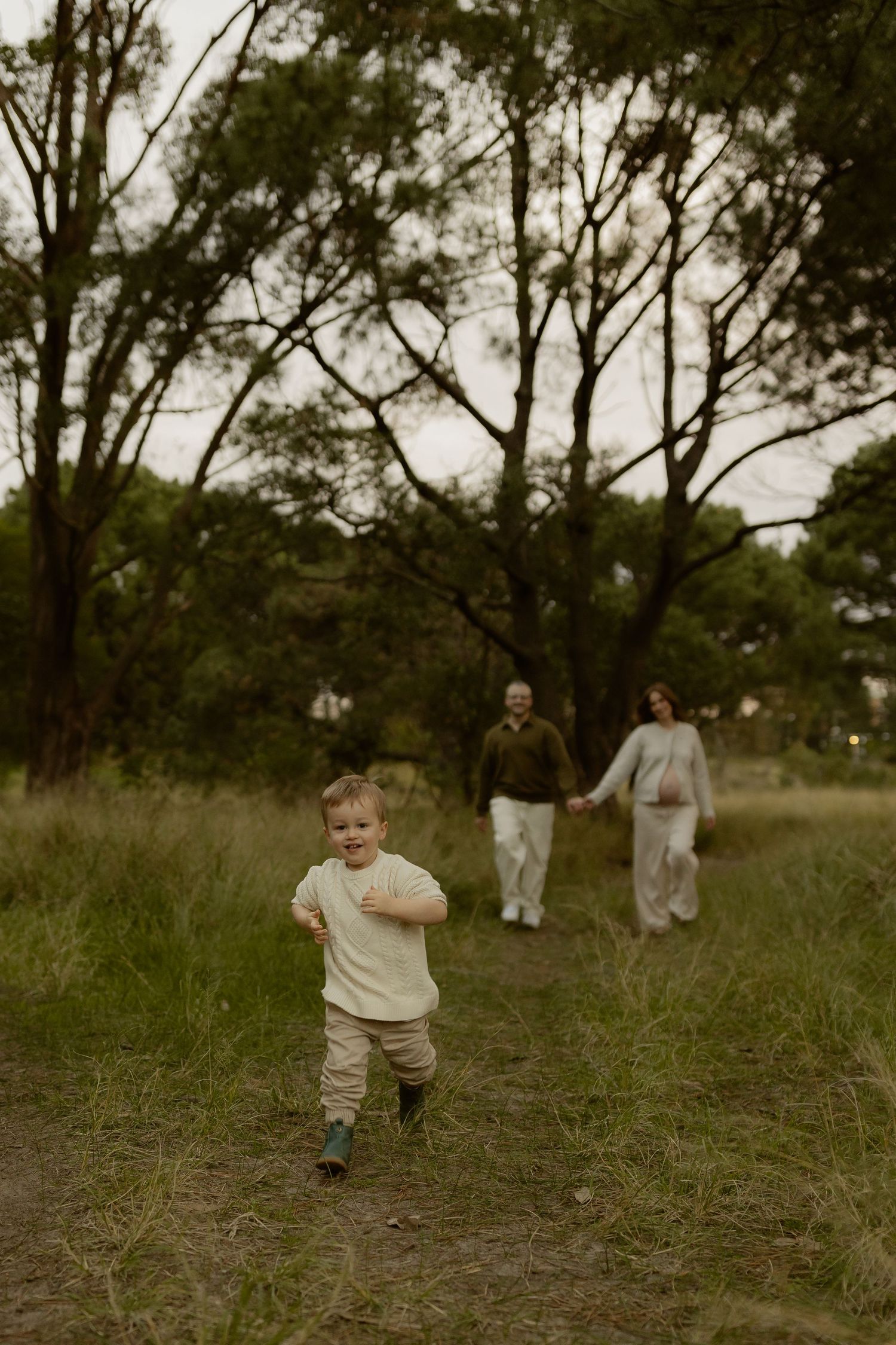 A toddler runs along a grassy path while his pregnant mother and father walk hand-in-hand behind him.