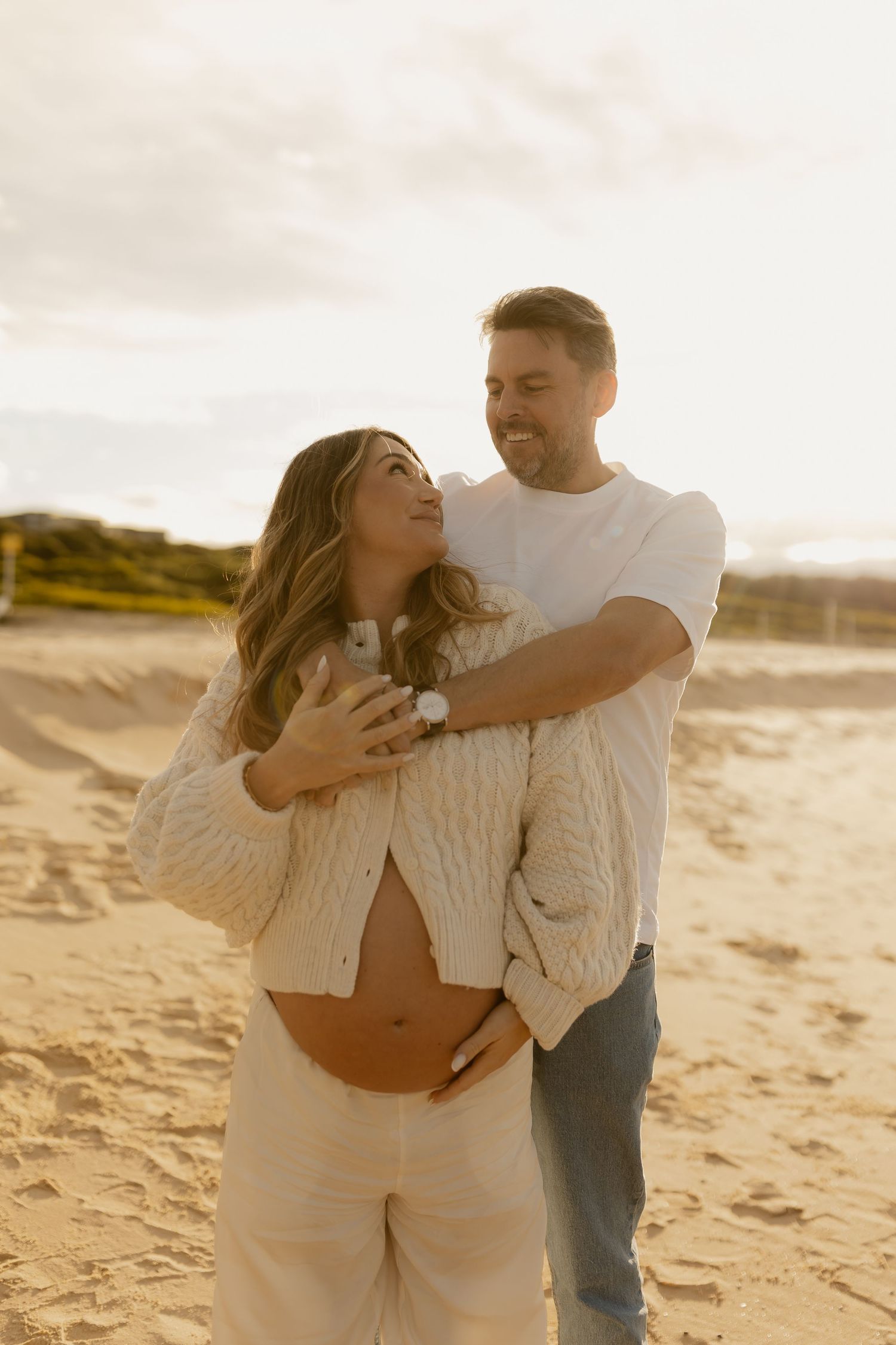 Happy couple embraces on golden sandy beach at sunset, man hugs pregnant woman from behind smiling.