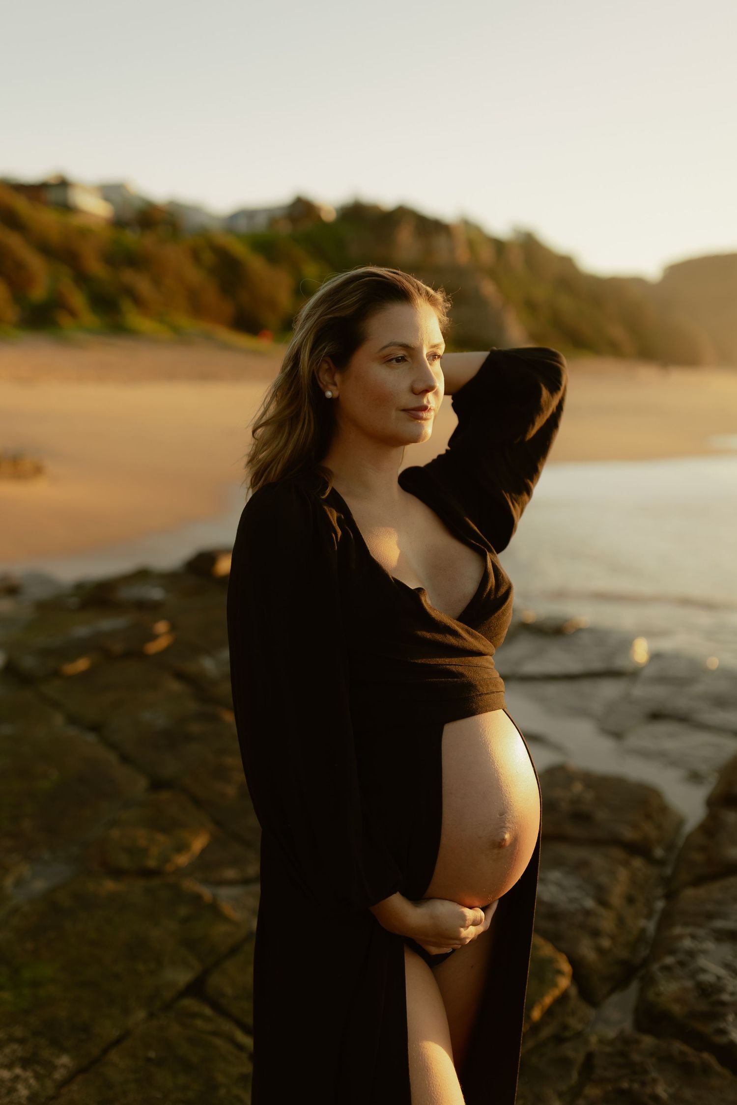 Pregnant woman in black wrap dress cradling baby bump at golden hour beach sunset.