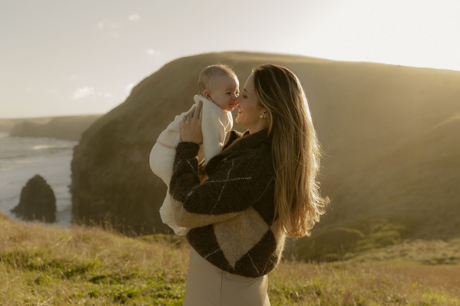 Mother kissing newborn baby on scenic coastal cliffs at golden hour.