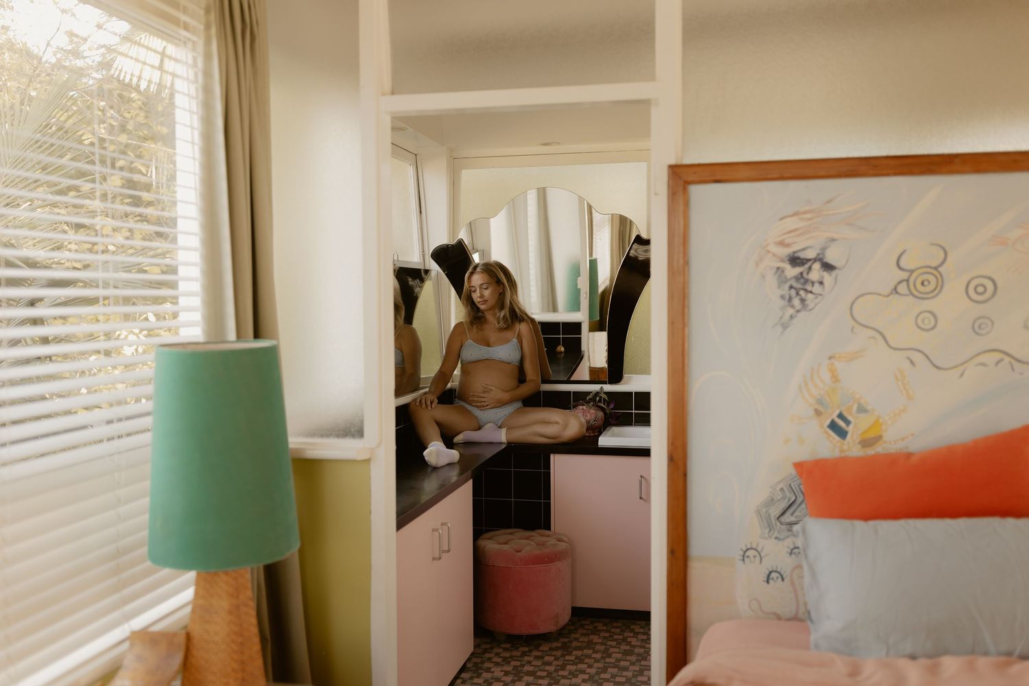 Woman sitting in a bright, stylish hotel room with a vanity mirror, teal lamp, and colorful wall art.