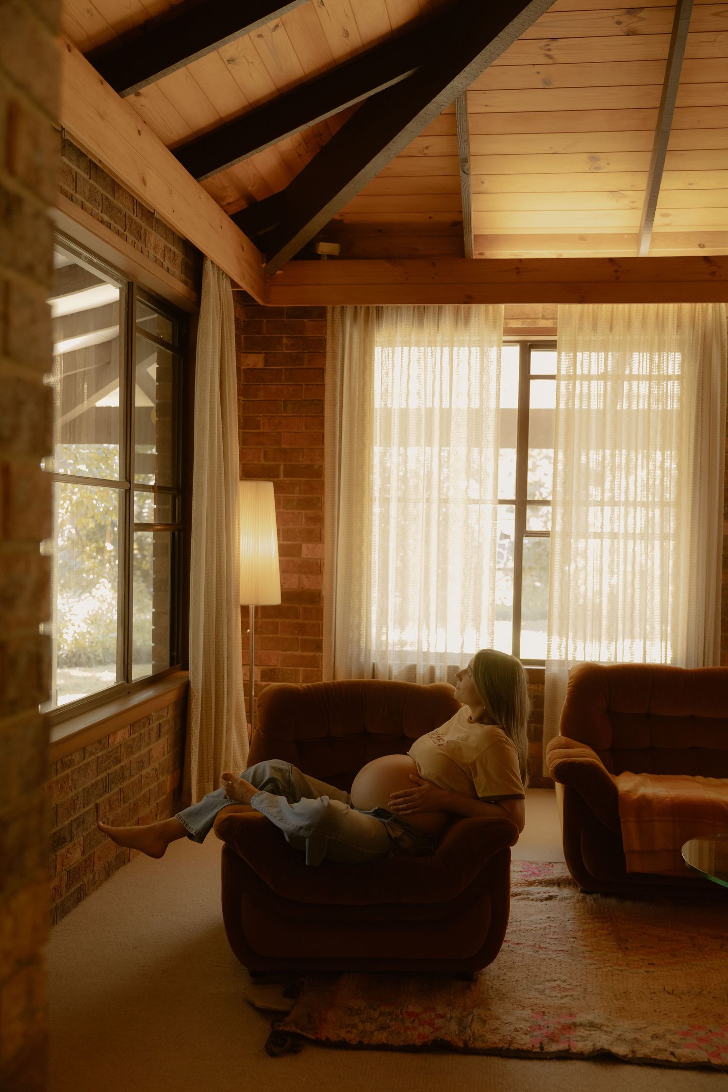 Pregnant woman relaxes alone in cozy brown armchair in rustic brick home with warm ambient lighting.