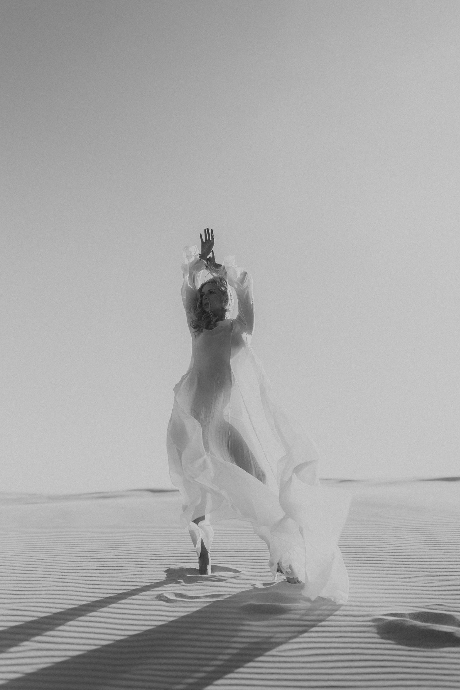 A woman in a flowing white dress dances gracefully on desert sand dunes in black and white.