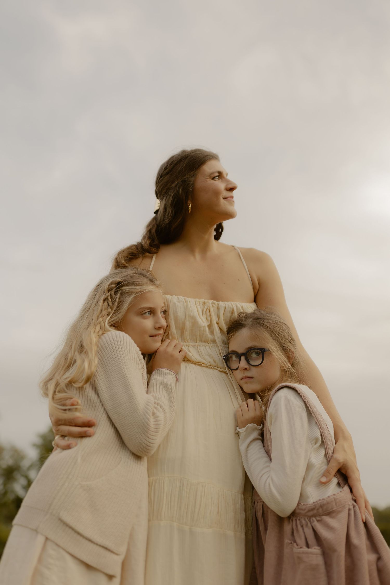 A woman in a white dress embraces two young girls outdoors under a cloudy sky in a serene portrait.